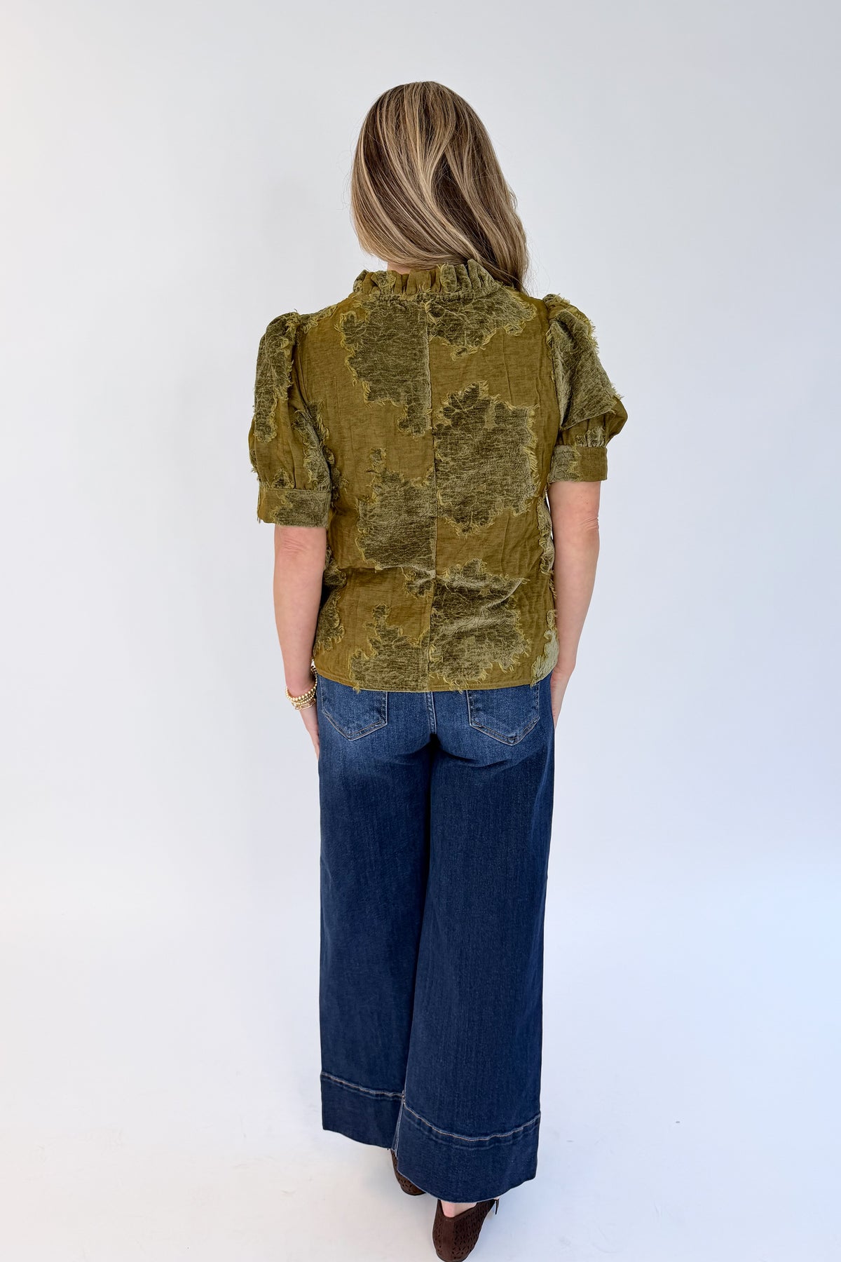 Back view of woman wearing Swagger Boutique’s Olive Velvet Ruffle Neck Top with jeans against white studio background.