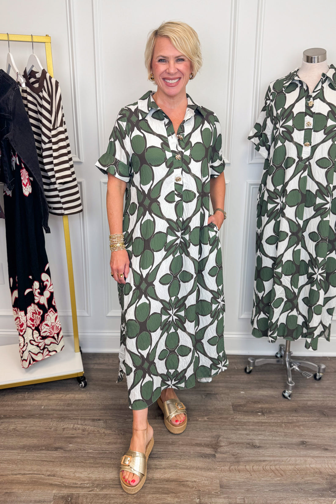 Front view of woman wearing Swagger Boutique’s Olive Abstract Print Midi Dress by Entro with sandals with clothing hanging in the background. 