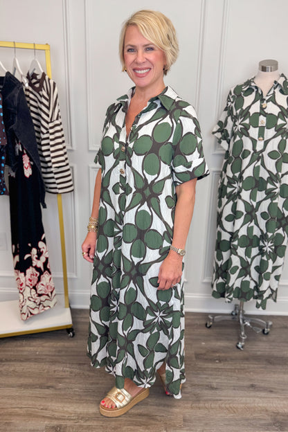 Side view of woman wearing Swagger Boutique’s Olive Abstract Print Midi Dress by Entro with sandals with clothing hanging in the background. 