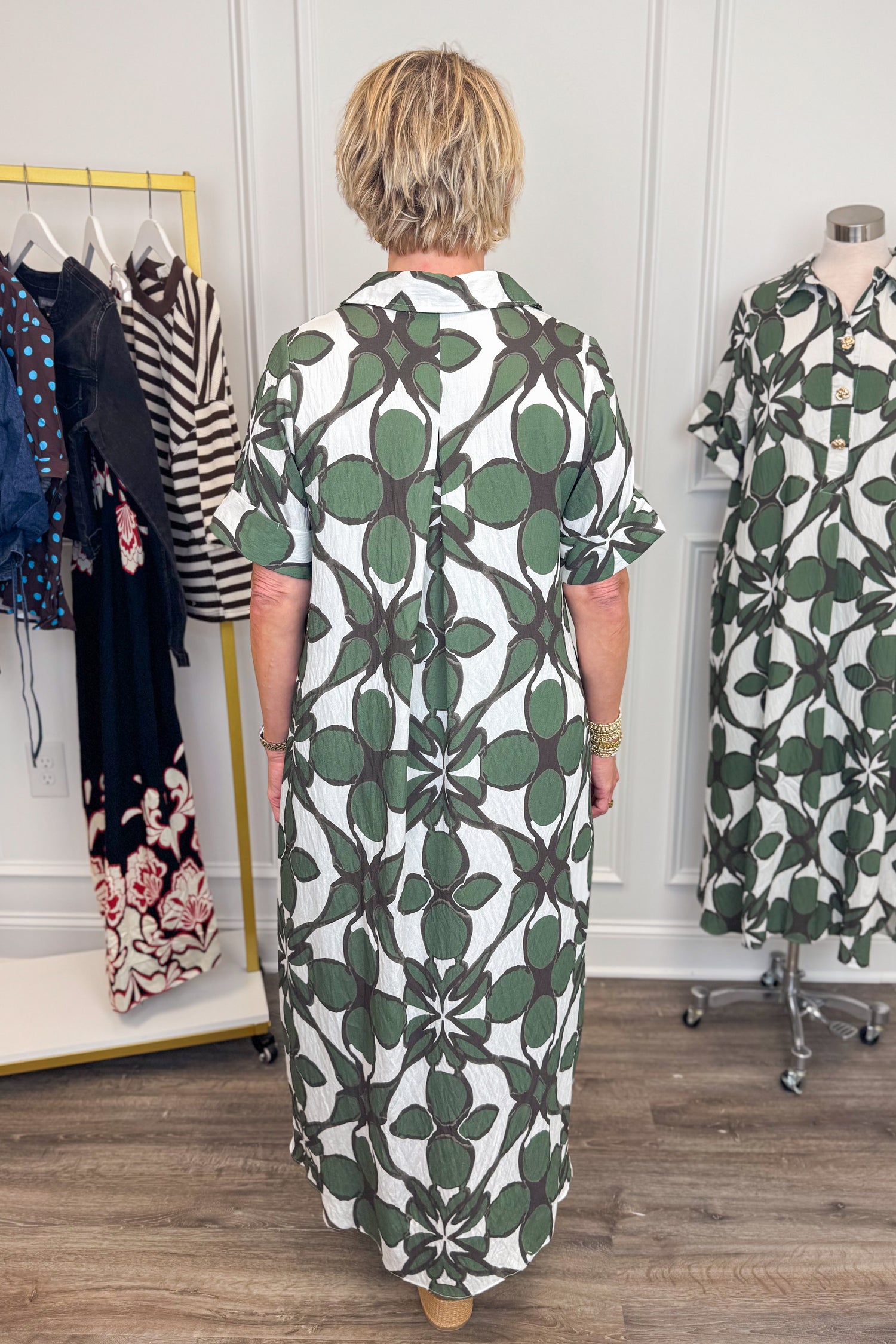 Back view of woman wearing Swagger Boutique’s Olive Abstract Print Midi Dress by Entro with sandals with clothing hanging in the background. 