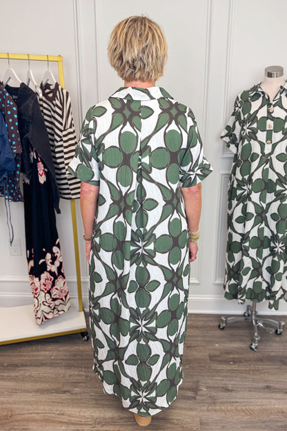 Back view of woman wearing Swagger Boutique’s Olive Abstract Print Midi Dress by Entro with sandals with clothing hanging in the background. 