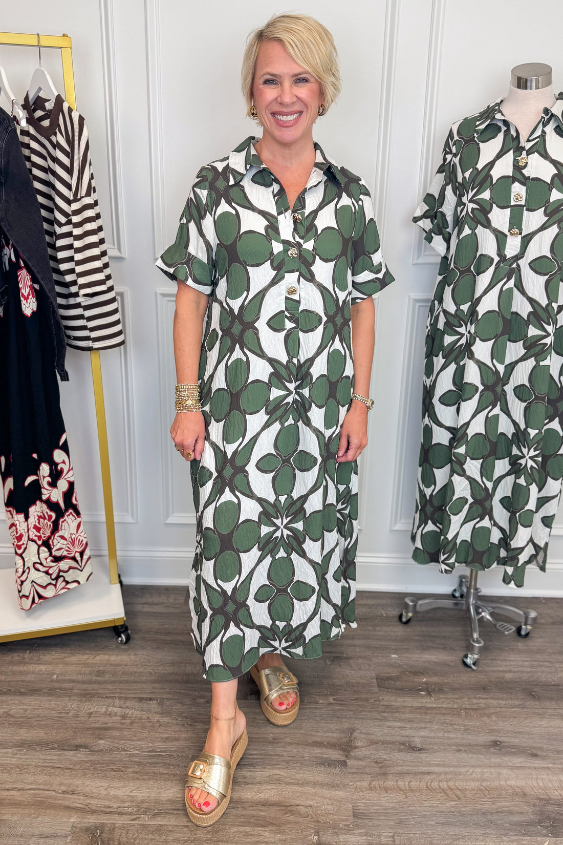 Front view of woman wearing Swagger Boutique’s Olive Abstract Print Midi Dress by Entro with sandals with clothing hanging in the background. 
