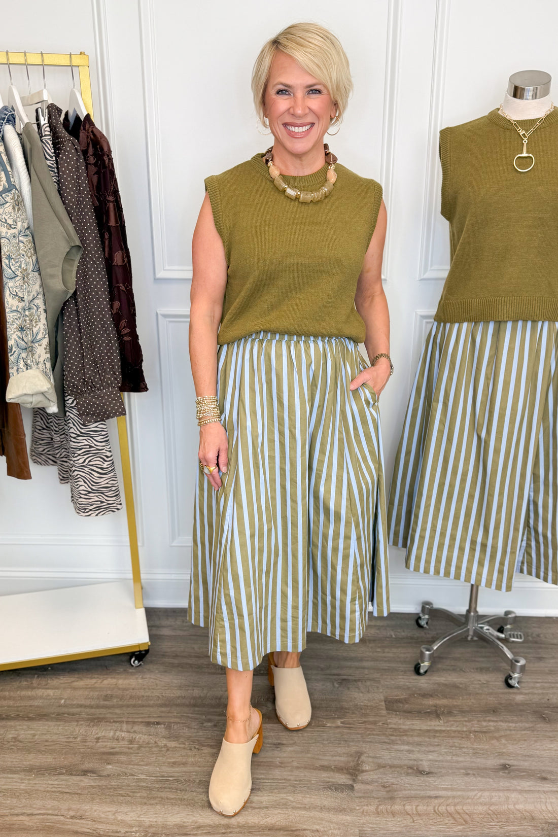 Front view of woman wearing Swagger Boutique’s Olive &amp; Blue Striped Midi Skirt with Olive Sleeveless Sweater with clothing hanging in the background.