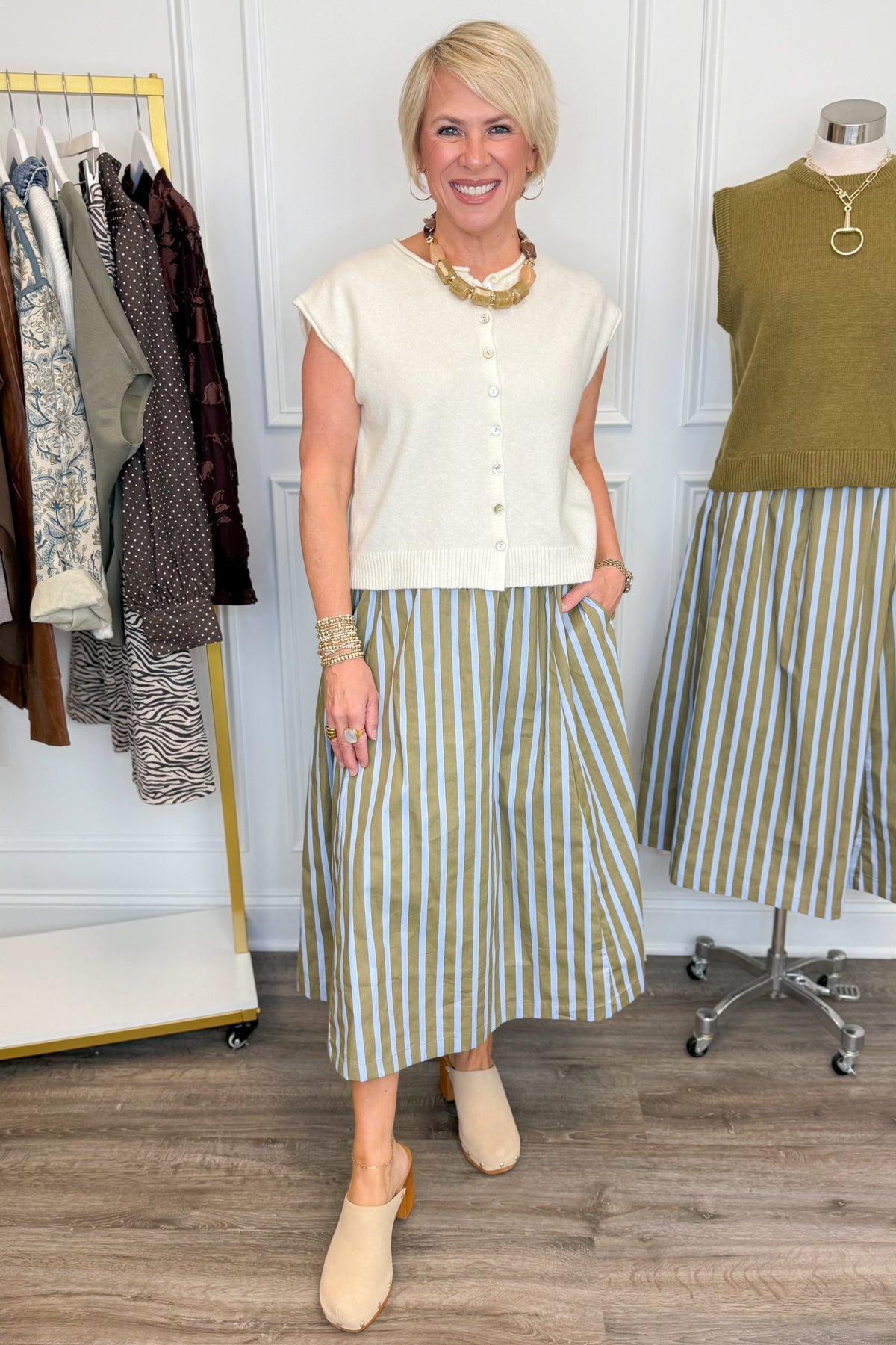 Front view of woman wearing Swagger Boutique’s Olive &amp; Blue Striped Midi Skirt with Cream Sleeveless Button Up Sweater with clothing hanging in the background.