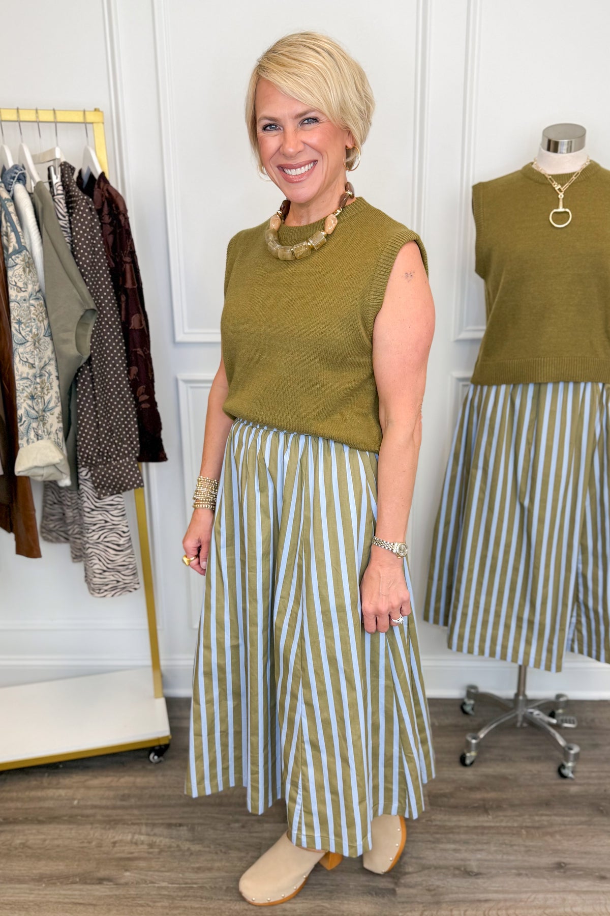 Side view of woman wearing Swagger Boutique’s Olive &amp; Blue Striped Midi Skirt with Olive Sleeveless Sweater with clothing hanging in the background.