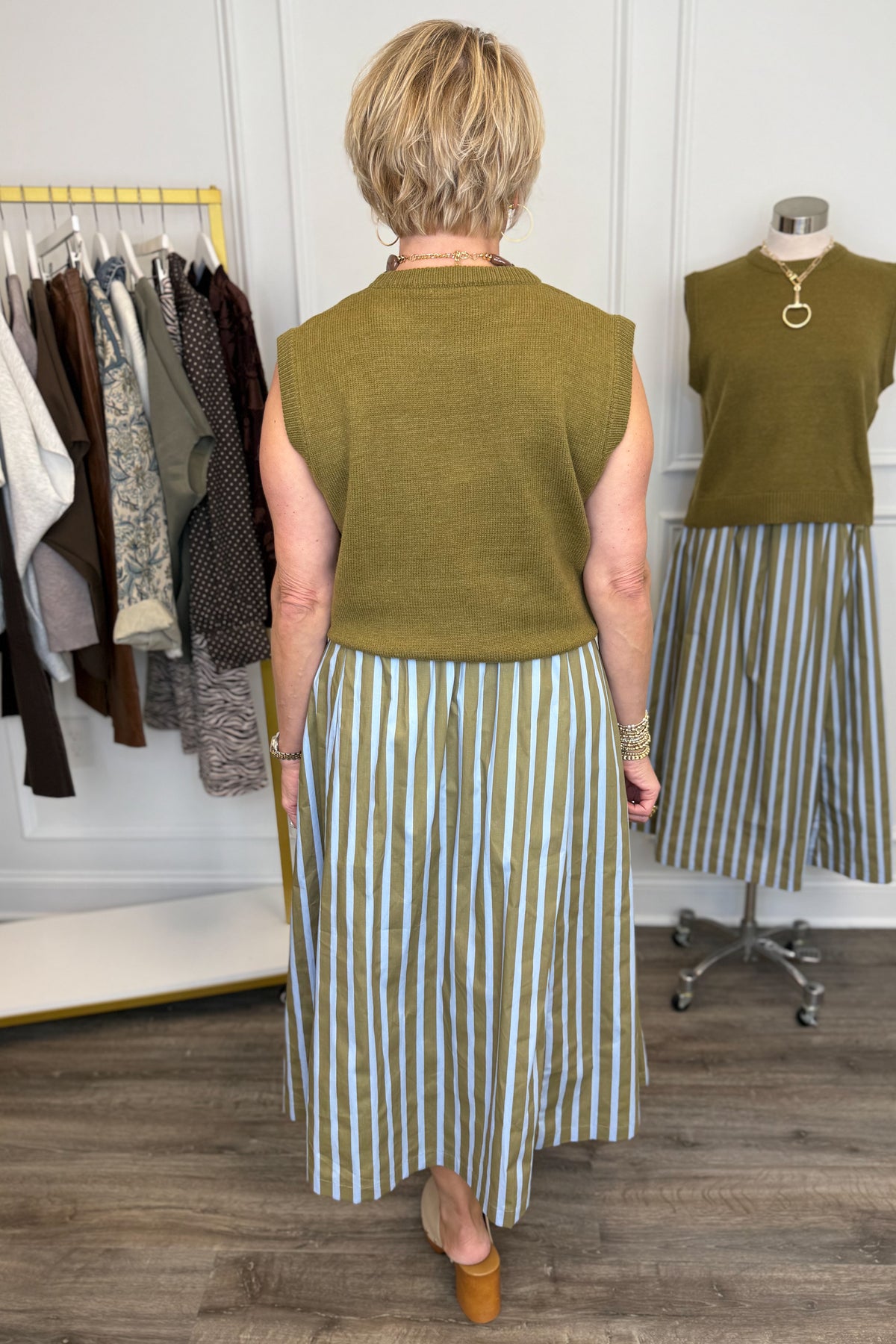 Back view of woman wearing Swagger Boutique’s Olive &amp; Blue Striped Midi Skirt with Olive Sleeveless Sweater with clothing hanging in the background.