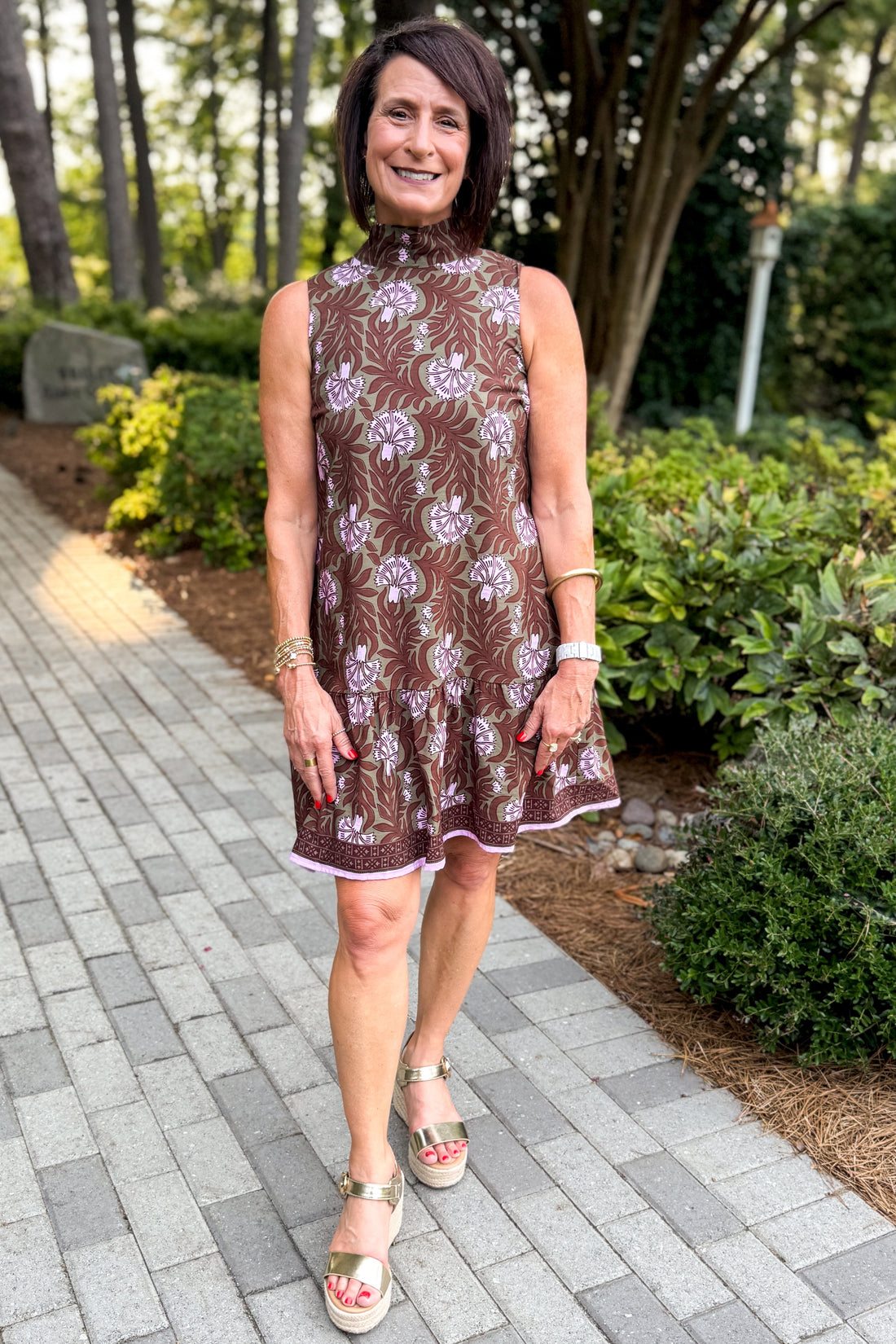 Front view of woman wearing Swagger Boutique’s Olive &amp; Brown Floral Halter Neck Mini Dress walking down a path outside.
