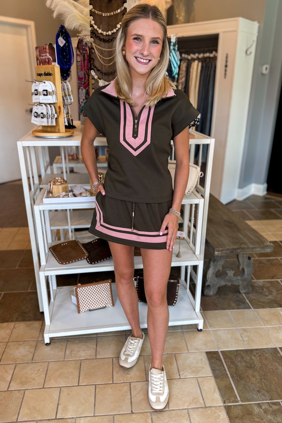 A woman modeling a dark brown short-sleeve dress with pink trim and a split neckline, standing in a boutique next to a white display shelf.