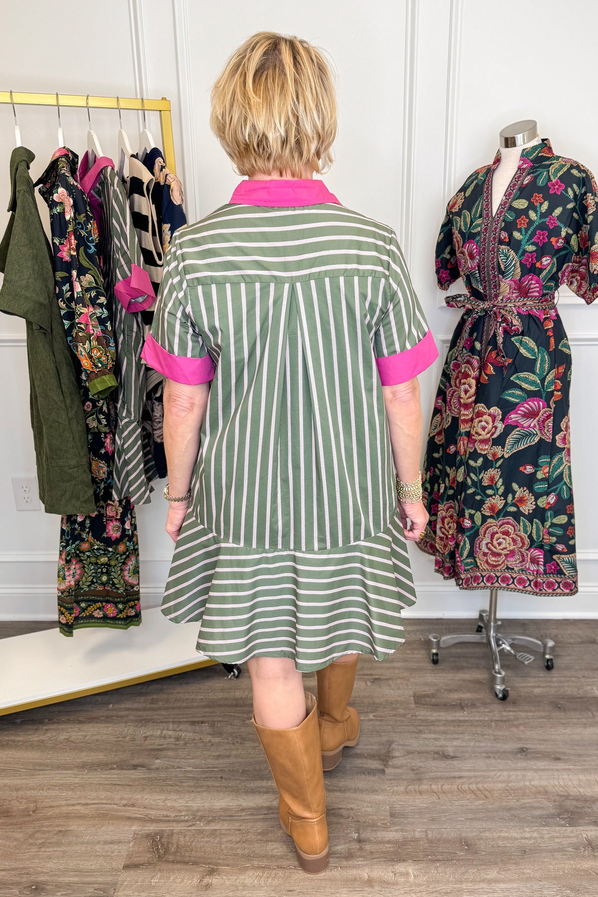 Back view of woman wearing Swagger Boutique’s Olive &amp; Pink Striped Mini Dress by Entro with boots with clothing hanging in the background.