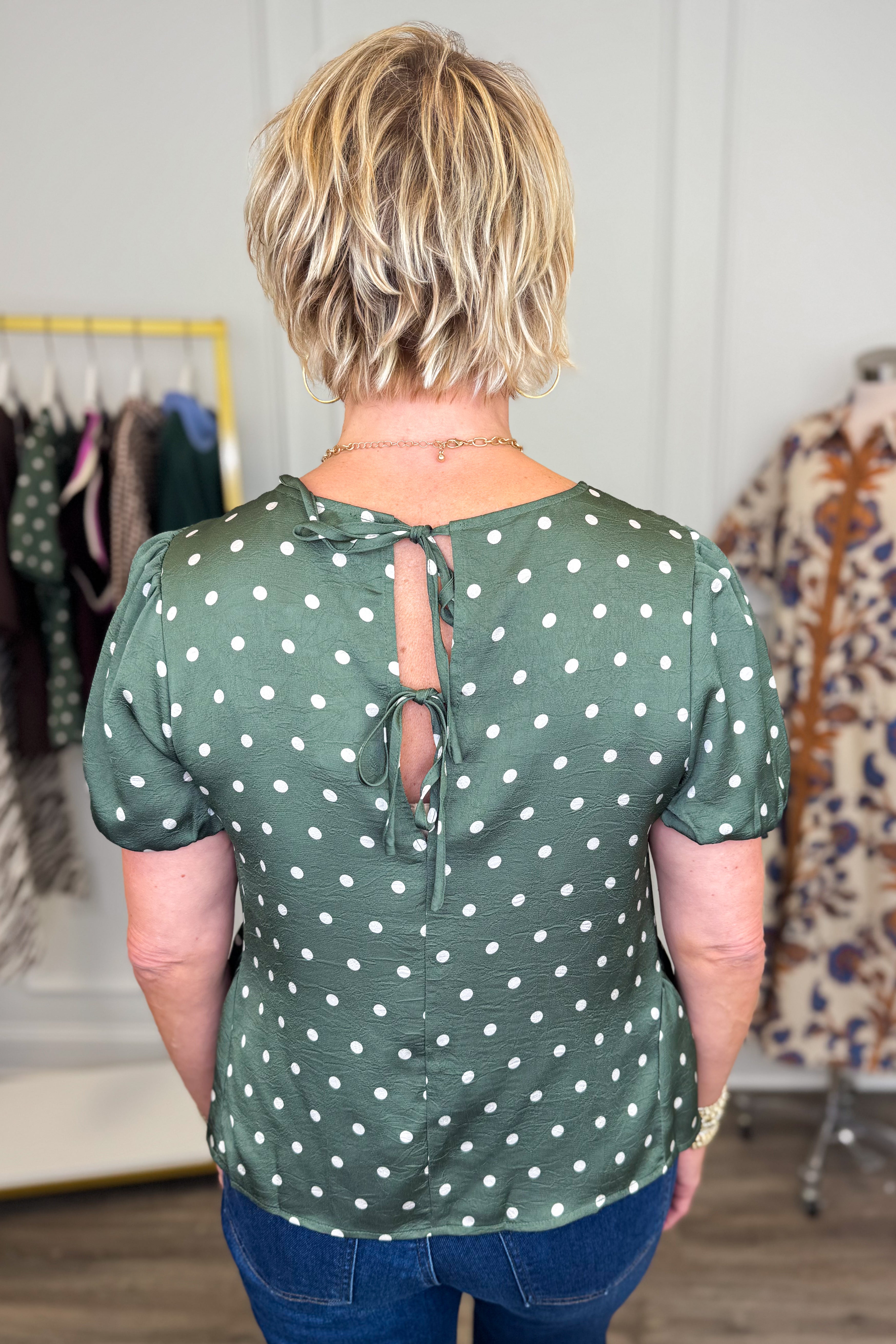 A woman viewed from the back wearing a sage green short-sleeve blouse with white polka dots and a tie keyhole back, paired with blue jeans in an indoor boutique setting.