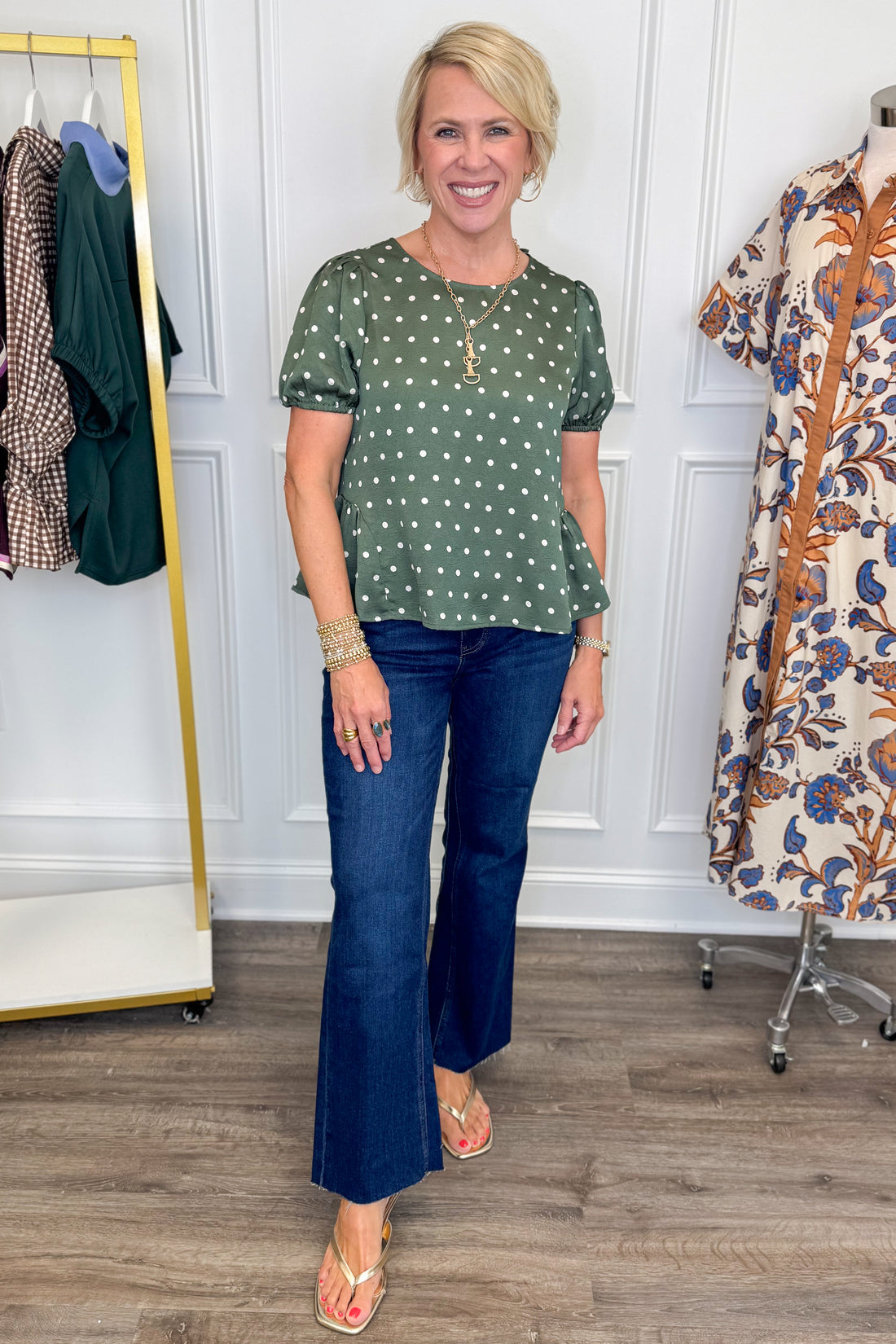 A woman modeling a green short-sleeve top with white polka dots and dark blue flare jeans, standing in a boutique setting with clothing racks visible in the background.