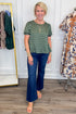 A woman modeling a green short-sleeve top with white polka dots and dark blue flare jeans, standing in a boutique setting with clothing racks visible in the background.