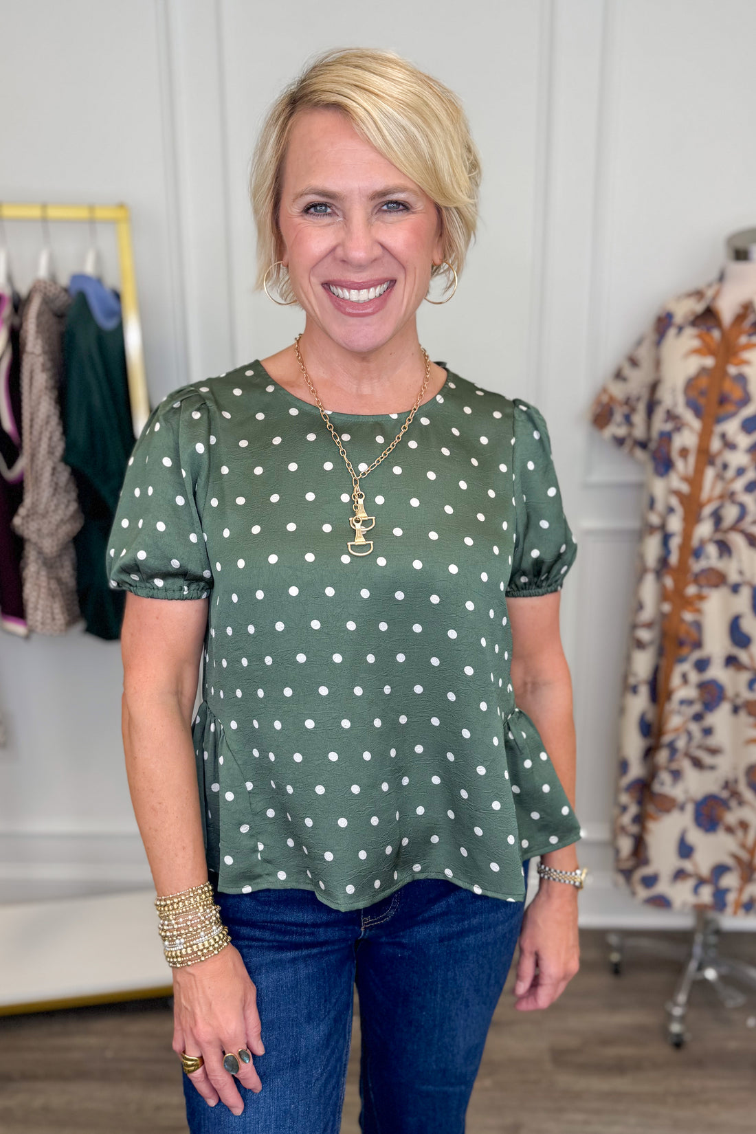 A smiling blonde woman wearing a sage green short-sleeve top with white polka dots and blue jeans, standing in a boutique setting with clothing racks visible in the background.