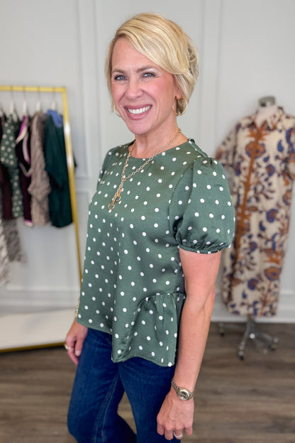 A smiling blonde woman wearing a sage green short-sleeve blouse with white polka dots and blue jeans, standing in a boutique setting with clothing racks and a mannequin in the background.