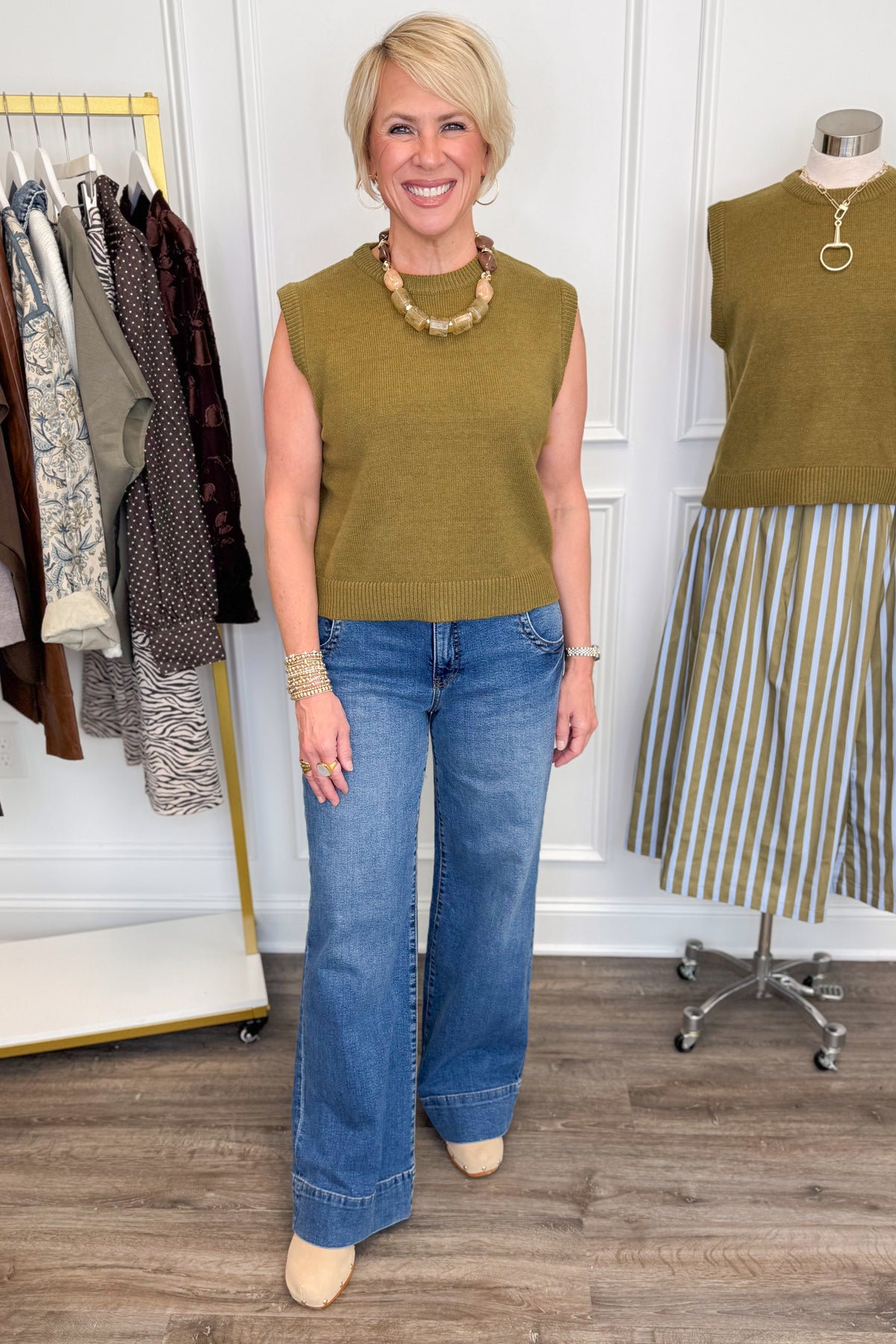 Front view of woman wearing Swagger Boutique’s Olive Sleeveless Sweater with jeans with clothing hanging in the background.
