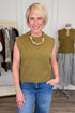 Front view of woman wearing Swagger Boutique’s Olive Sleeveless Sweater with jeans with clothing hanging in the background.