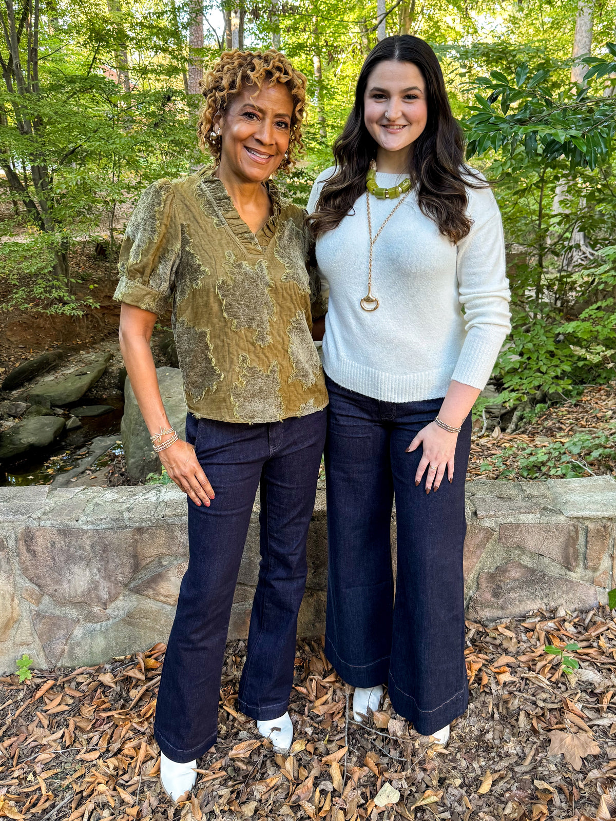 Front view of two women wearing Swagger Boutique’s Olive Velvet Ruffle Neck Top and Ivory Ribbed Hem Sweater with jeans outside at a park.