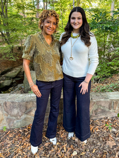 Front view of two women wearing Swagger Boutique’s Olive Velvet Ruffle Neck Top and Ivory Ribbed Hem Sweater with jeans outside at a park.