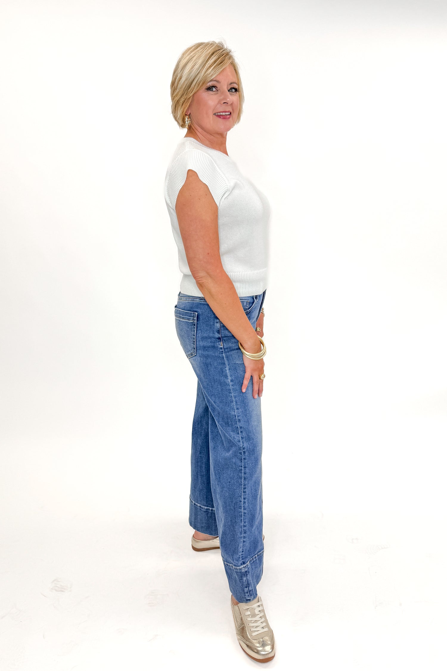 Side view of woman wearing Medium Wash Pintuck Detail Jeans by Risen with white sweater against white studio background.