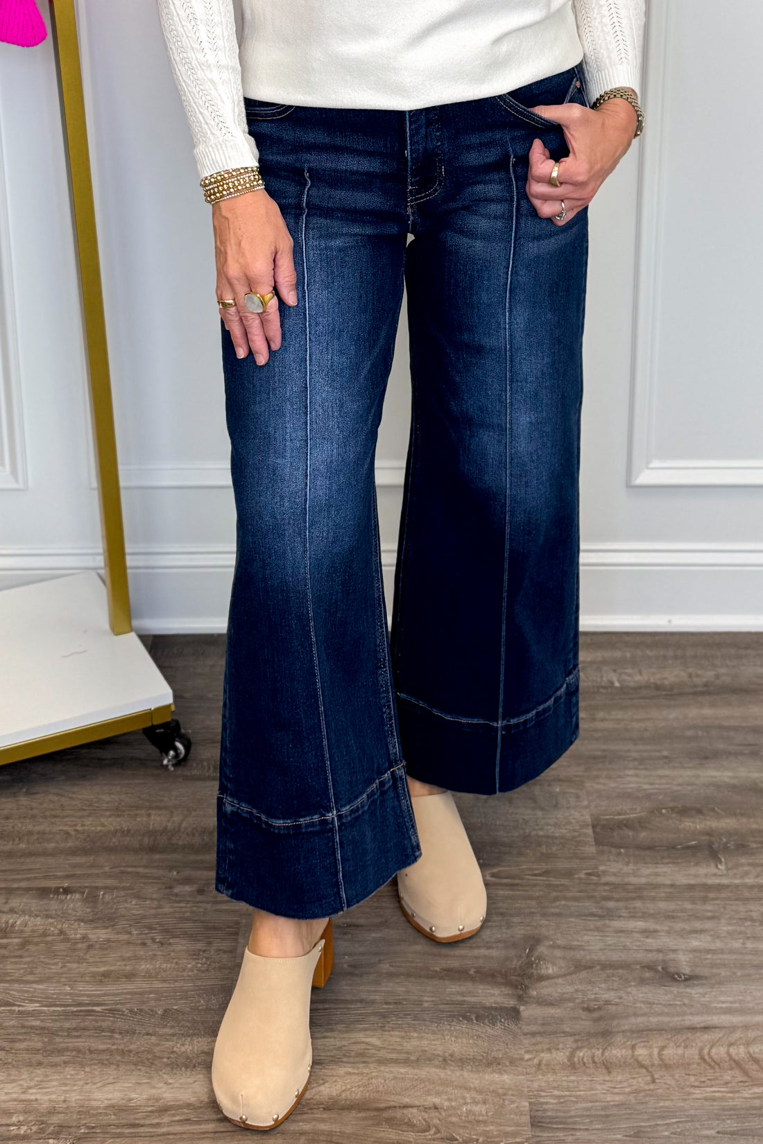 Front view of woman wearing Swagger Boutique’s sweater with Parker Pintuck Dark Wash Denim Jeans by Risen with clothing hanging in the background.