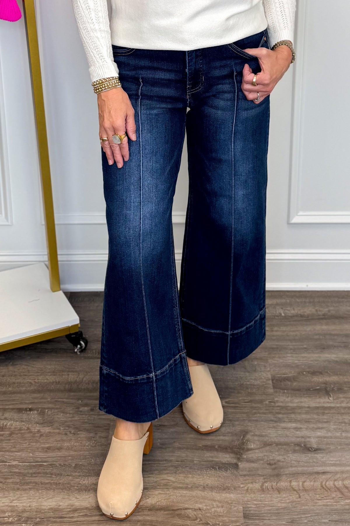 Front view of woman wearing Swagger Boutique’s sweater with Parker Pintuck Dark Wash Denim Jeans by Risen with clothing hanging in the background.