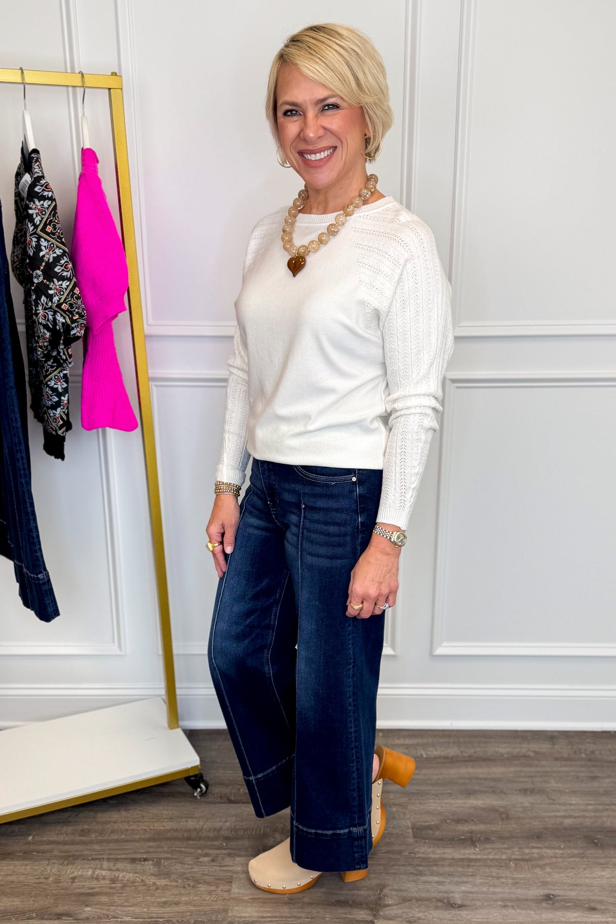 Side view of woman wearing Swagger Boutique’s sweater with Parker Pintuck Dark Wash Denim Jeans by Risen with clothing hanging in the background.