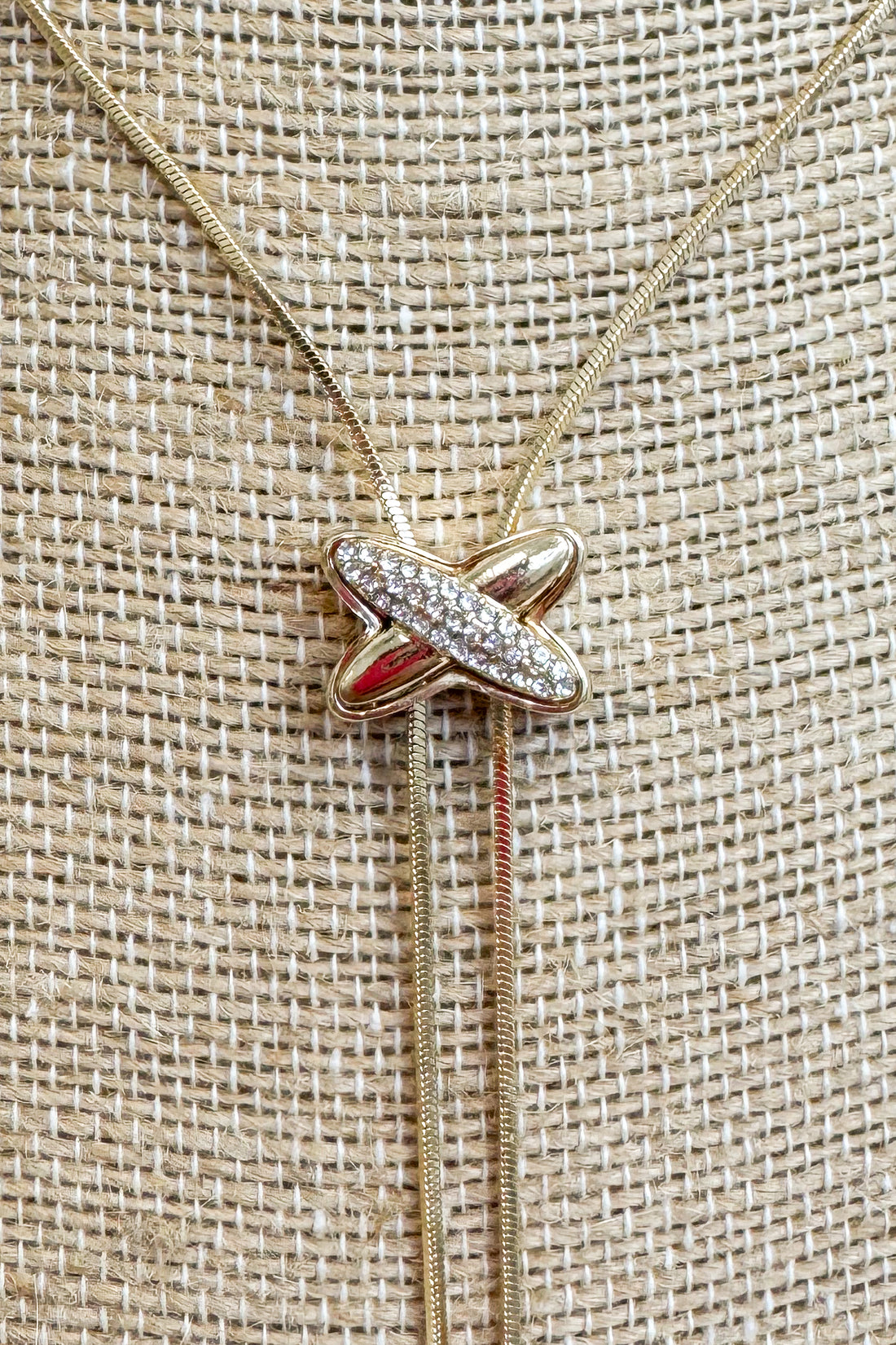 Close up of Gold-tone delicate chain necklace featuring a elongated vertical X pendant on a beige jewelry bust