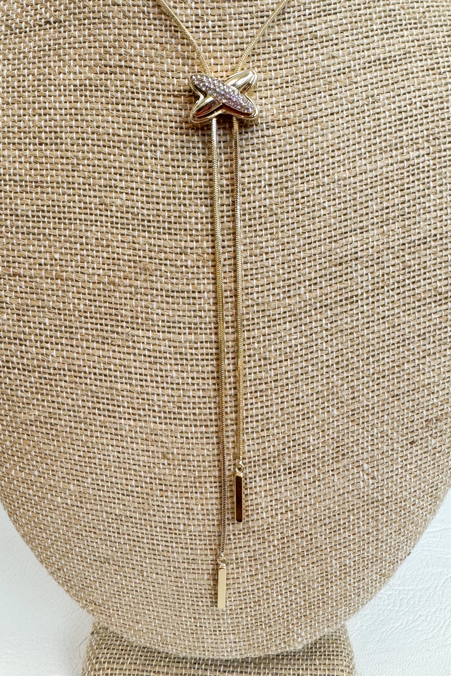 Close up of Gold-tone delicate necklace featuring a elongated vertical X pendant with two gold dangles on a beige jewelry bust