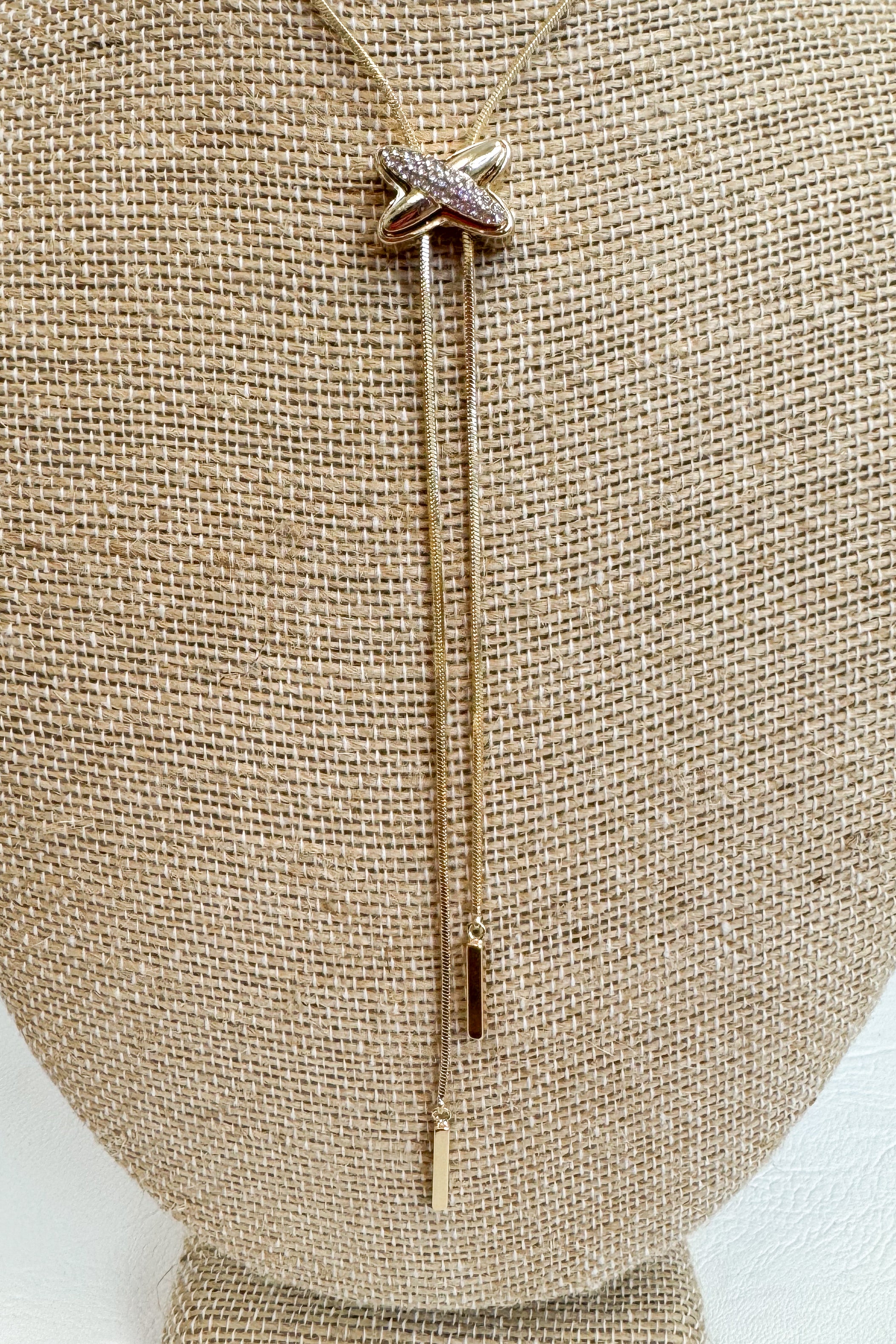 Close up of Gold-tone delicate necklace featuring a elongated vertical X pendant with two gold dangles on a beige jewelry bust