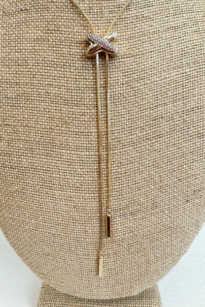 Close up of Gold-tone delicate necklace featuring a elongated vertical X pendant with two gold dangles on a beige jewelry bust