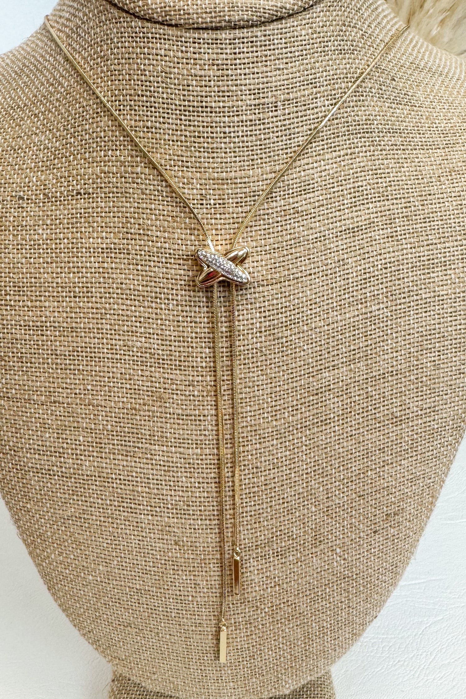  Gold-tone delicate necklace featuring a elongated vertical X pendant on a beige jewelry bust