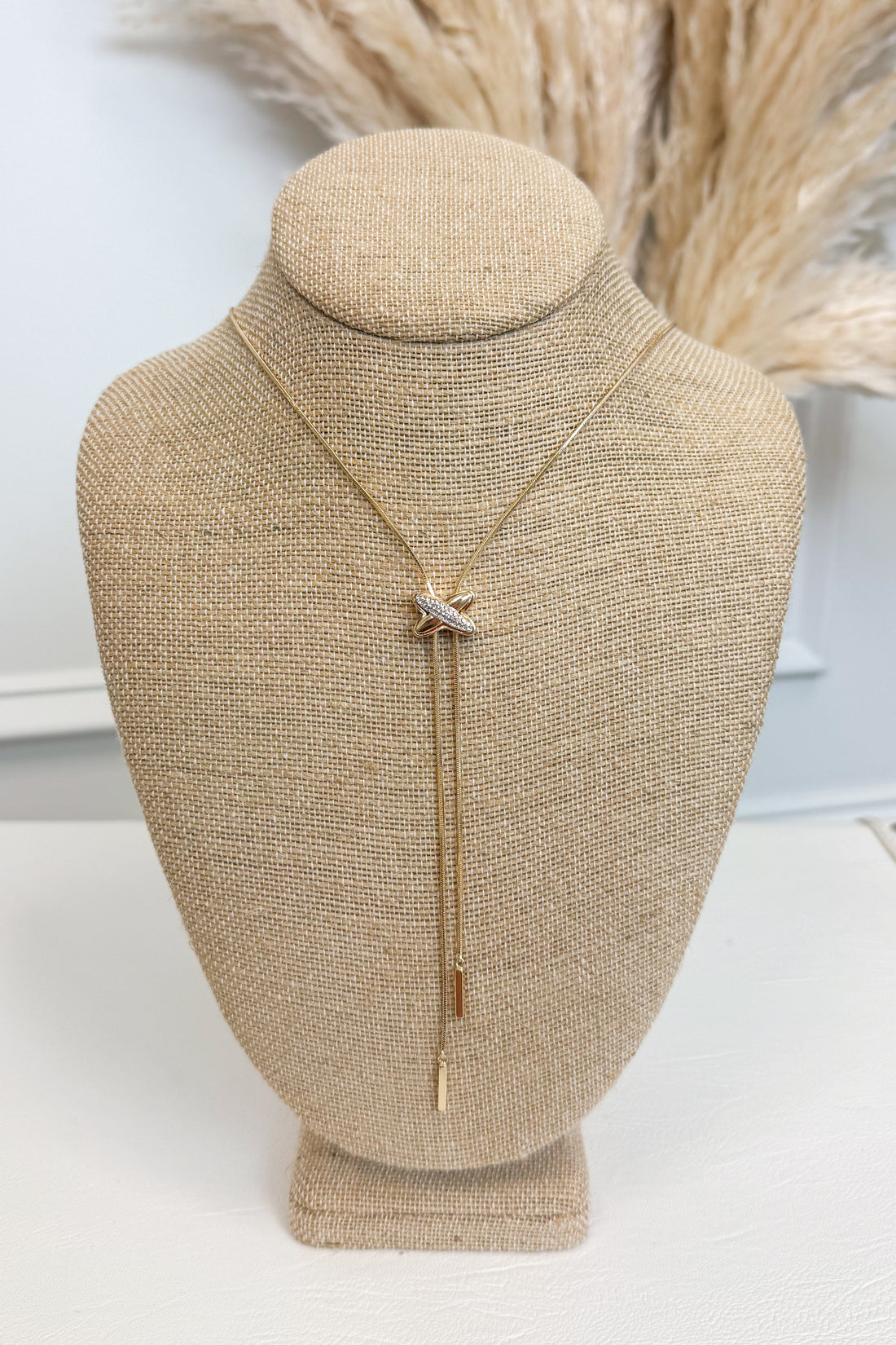 Gold-tone delicate chain necklace featuring a small elongated vertical X pendant on a beige jewelry bust