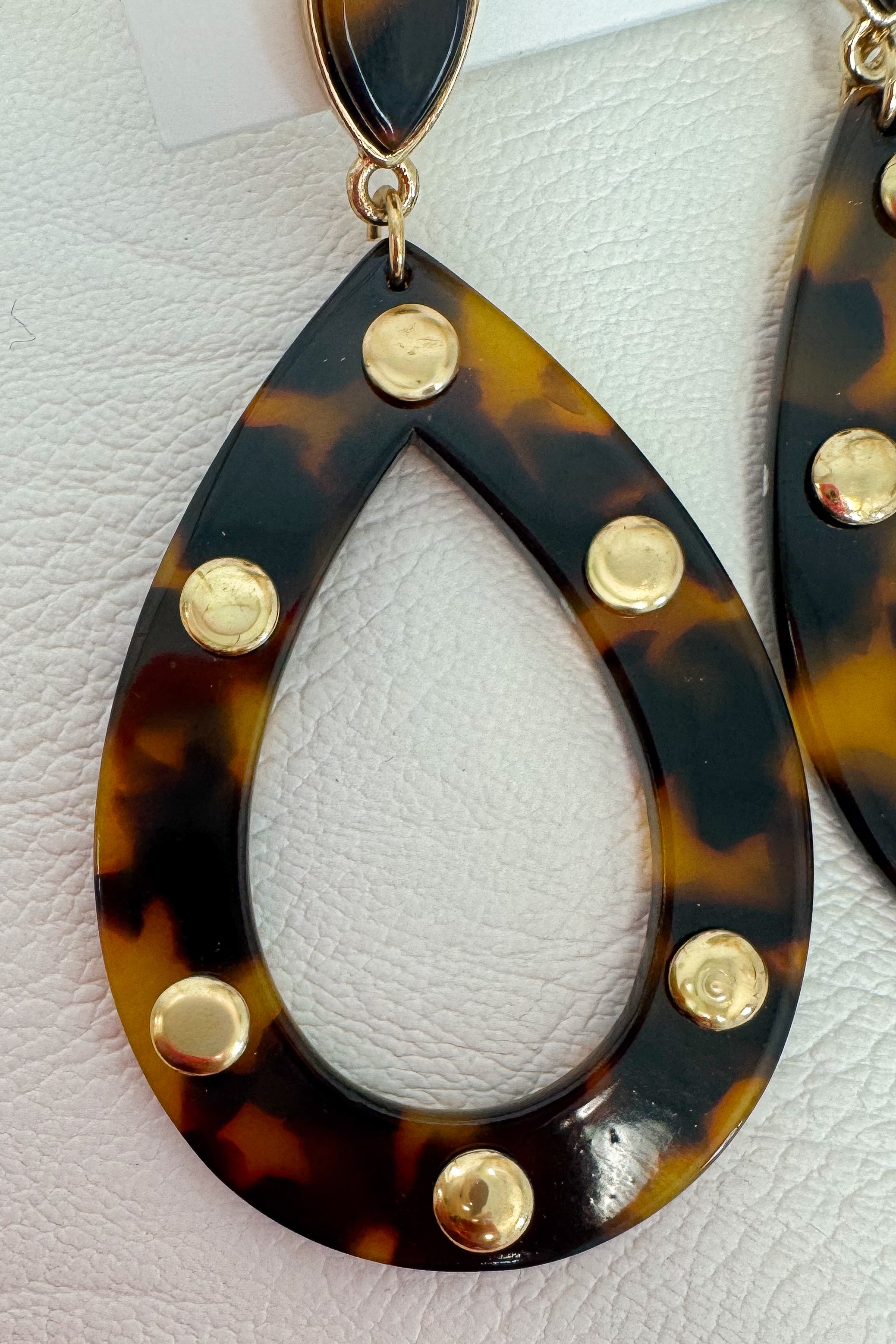 Close-up of a teardrop-shaped dangling earring with a glossy tortoiseshell pattern in dark brown and amber, accented with gold-tone metal studs and a gold top loop, shown against a light textured background.