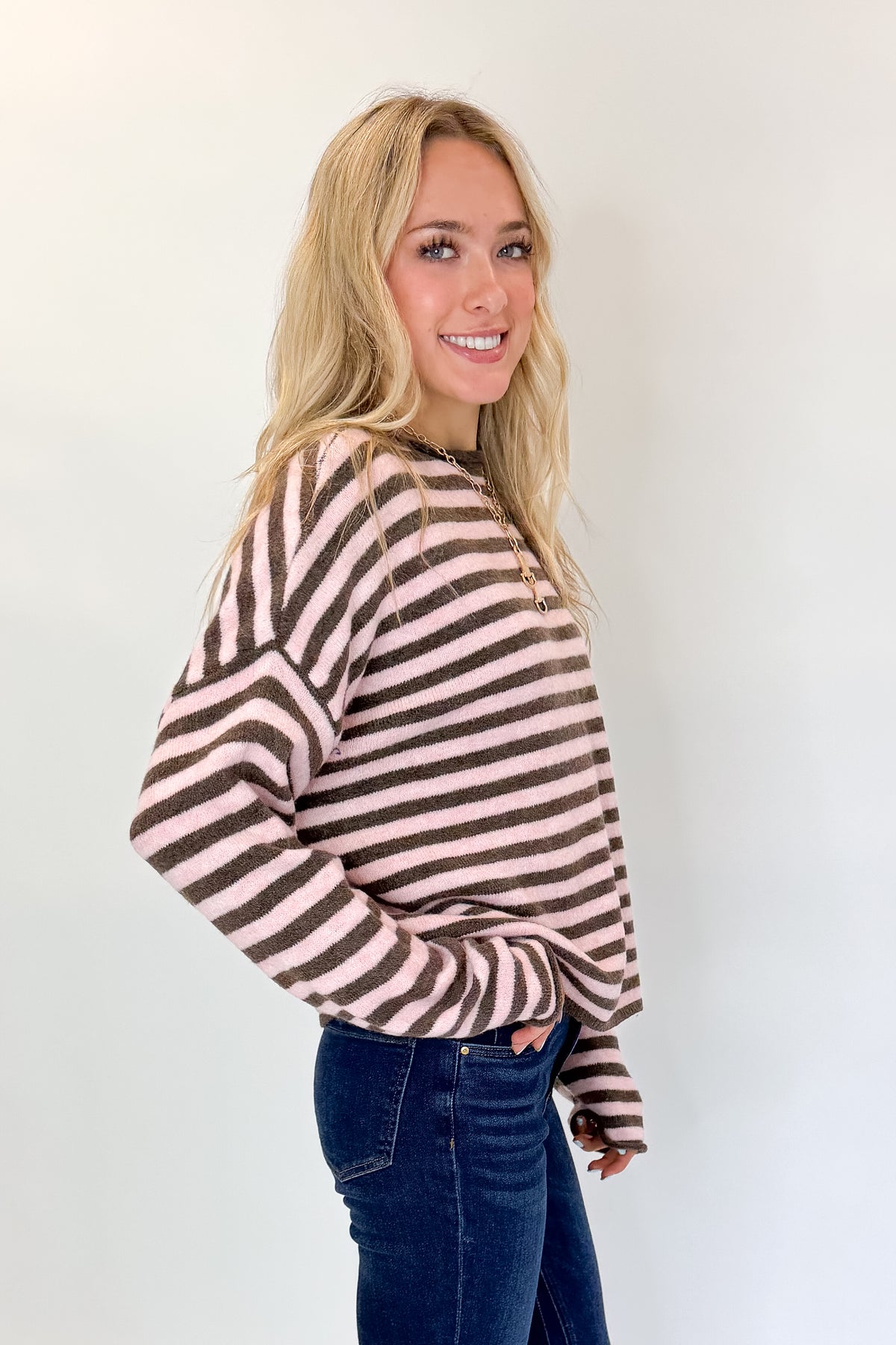 Side view of woman wearing Swagger Boutique’s Pink &amp; Brown Striped Sweater with jeans against white studio background.