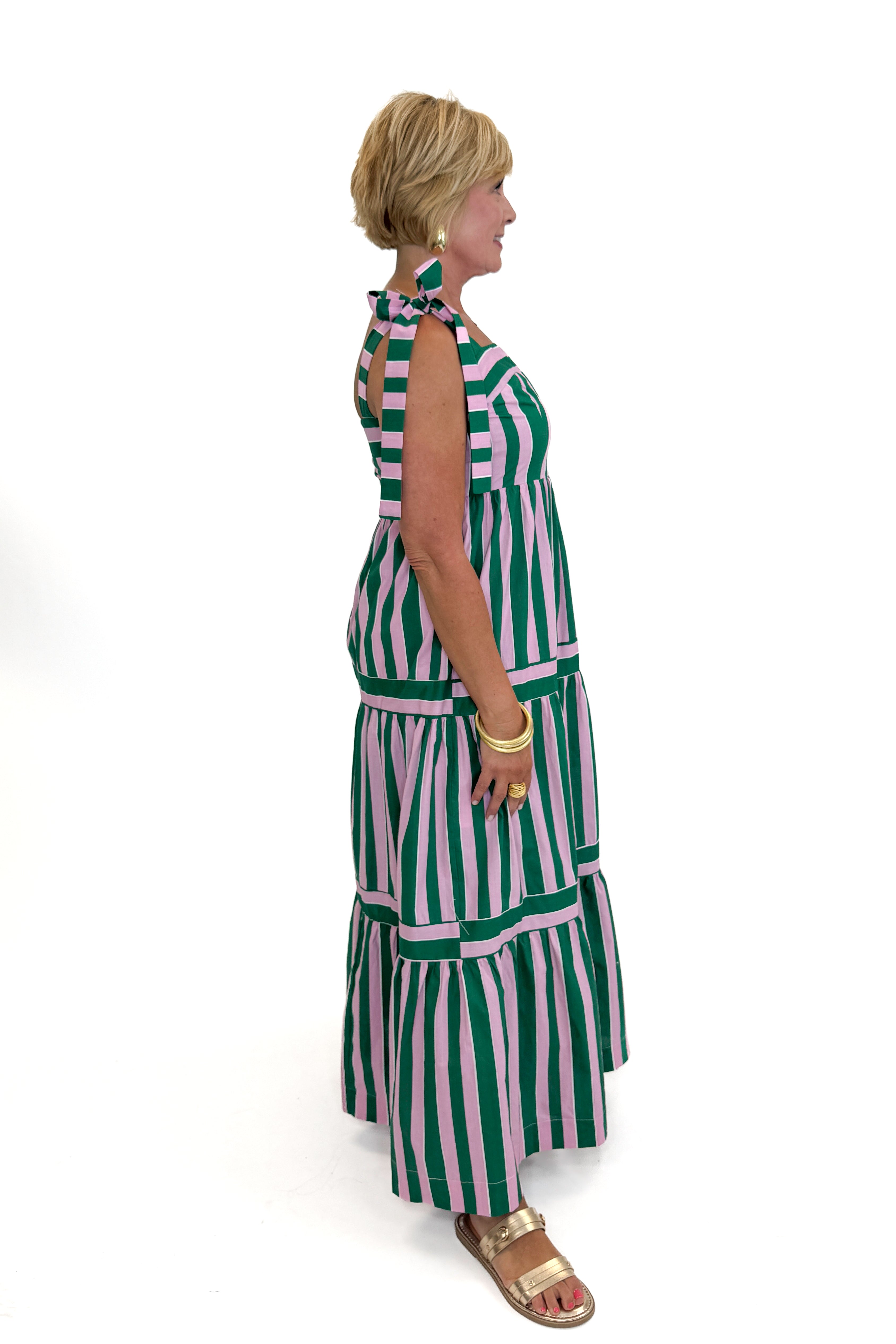 Side view of woman wearing Swagger Boutique’s Pink &amp; Green Striped Tiered Maxi Dress with sandals against white studio background.