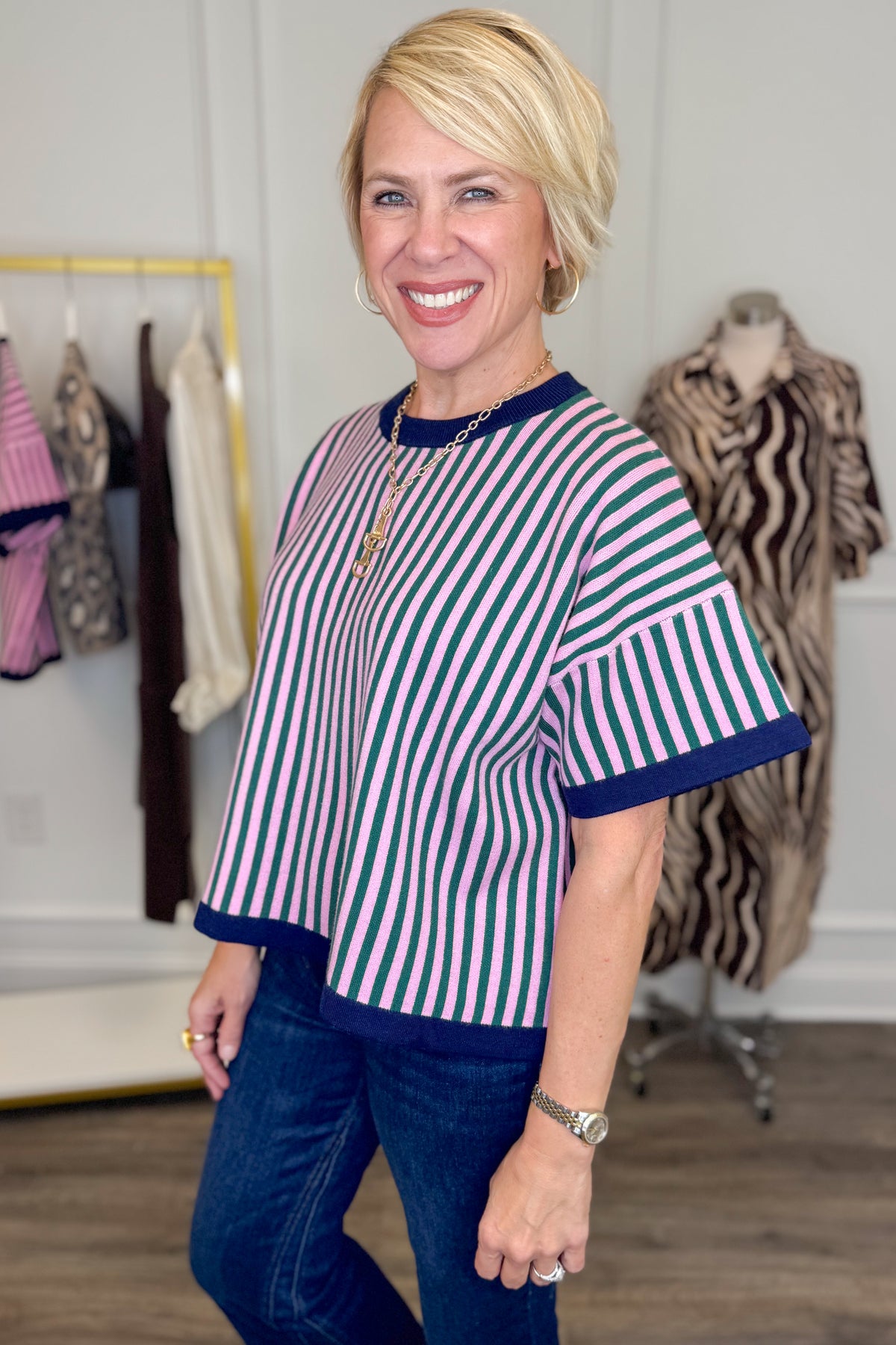 Side view of woman wearing Swagger Boutique’s Pink &amp; Green Striped Short Sleeve Sweater with jeans with clothing hanging in the background.