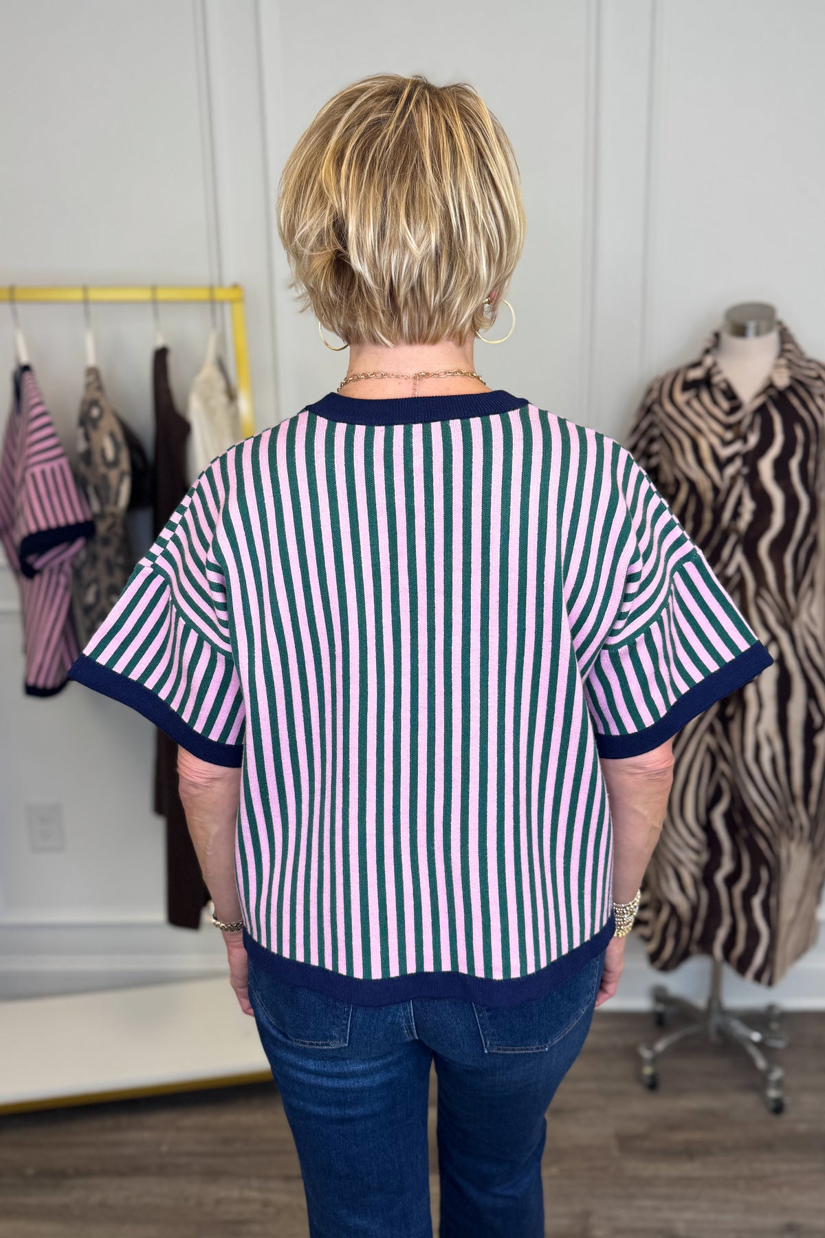 Back view of woman wearing Swagger Boutique’s Pink &amp; Green Striped Short Sleeve Sweater with jeans with clothing hanging in the background.