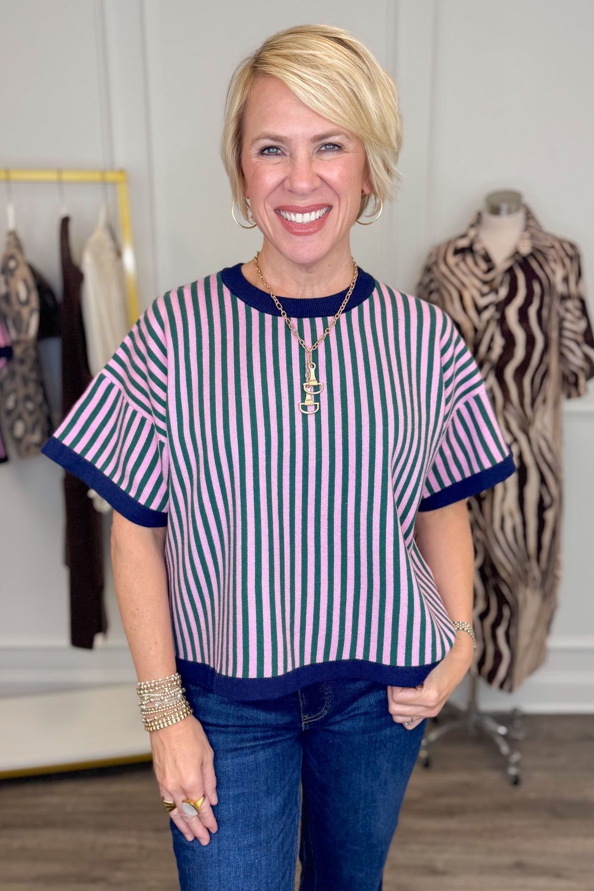 Front view of woman wearing Swagger Boutique’s Pink &amp; Green Striped Short Sleeve Sweater with jeans with clothing hanging in the background.