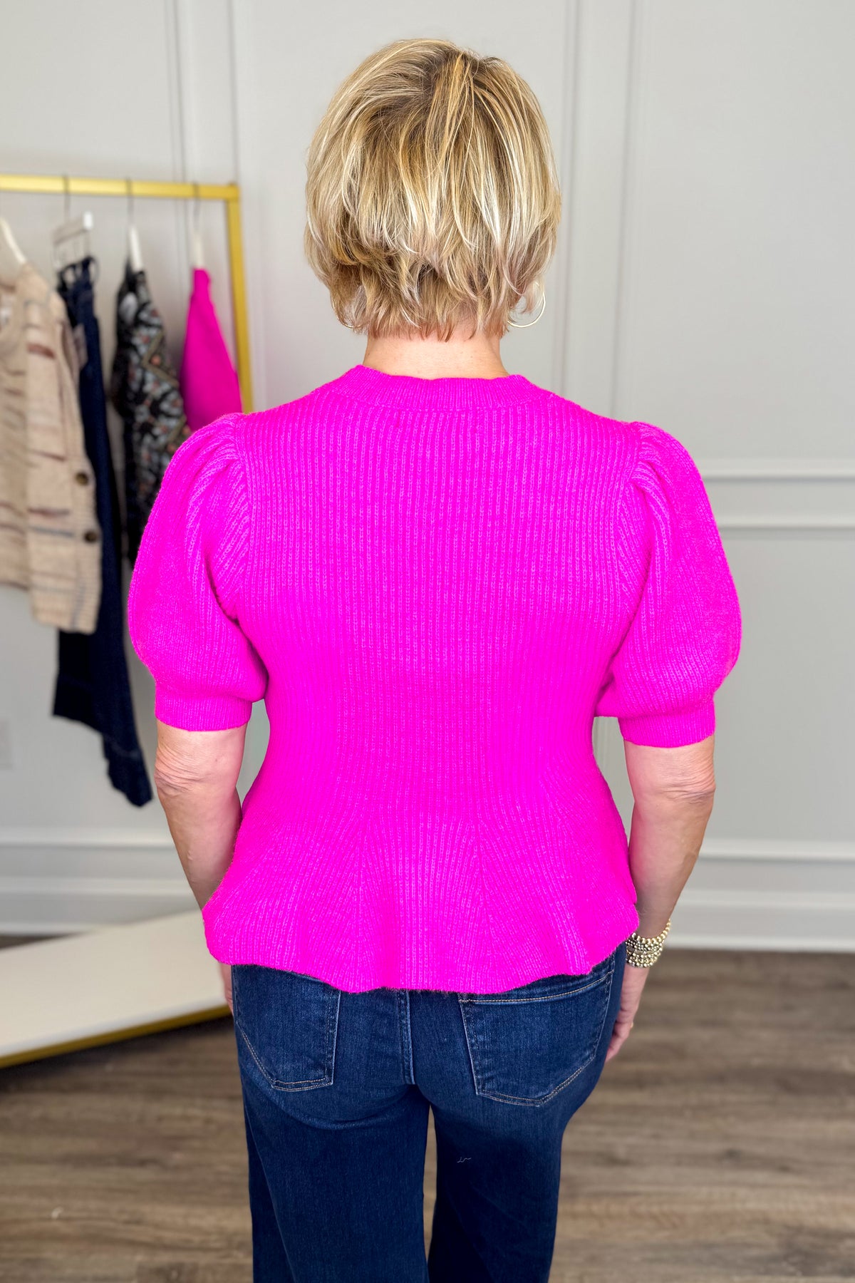 Back view of woman wearing Swagger Boutique’s Pink Peplum Short Sleeve Sweater with jeans with clothing hanging in the background.