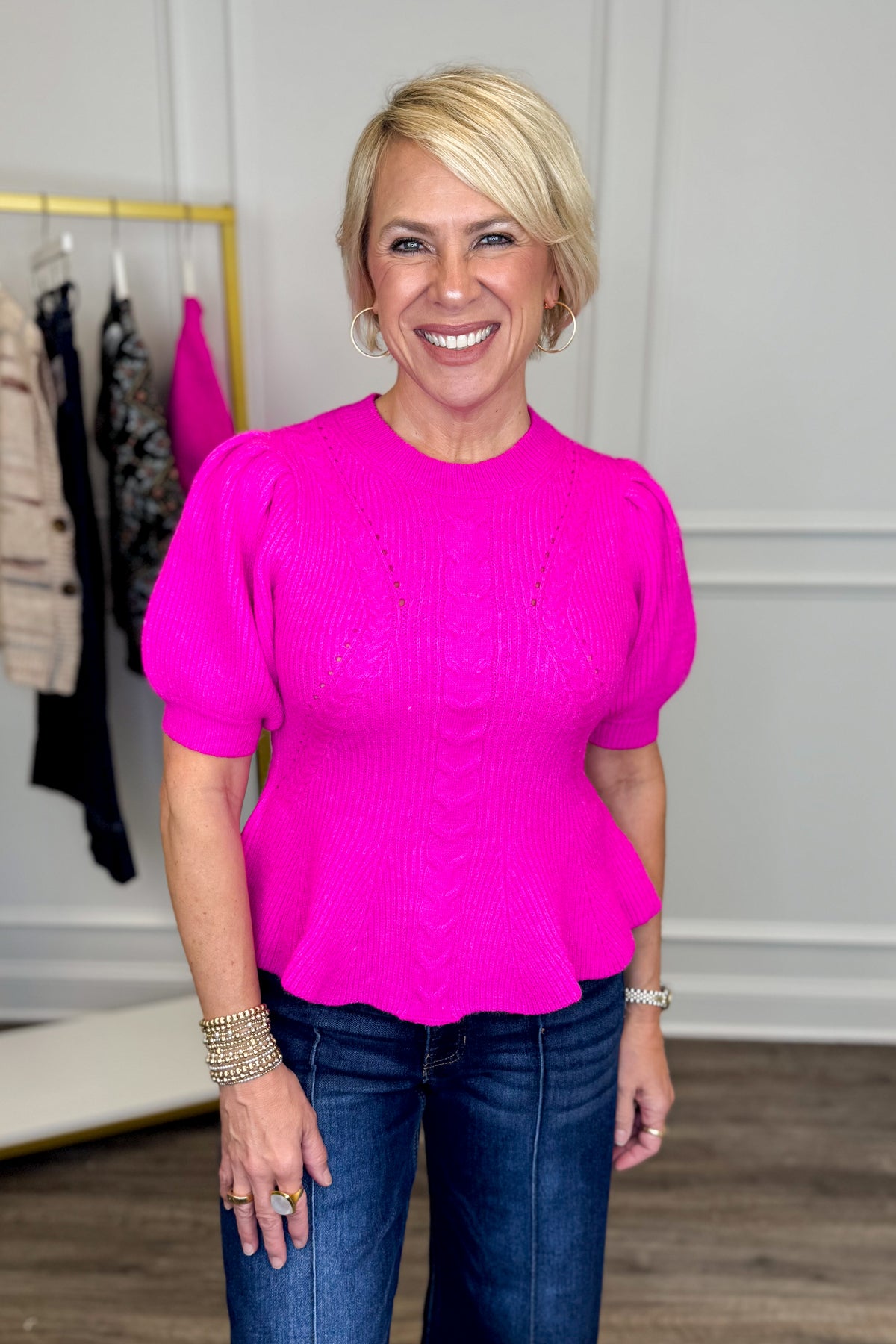 Front view of woman wearing Swagger Boutique’s Pink Peplum Short Sleeve Sweater with jeans with clothing hanging in the background.