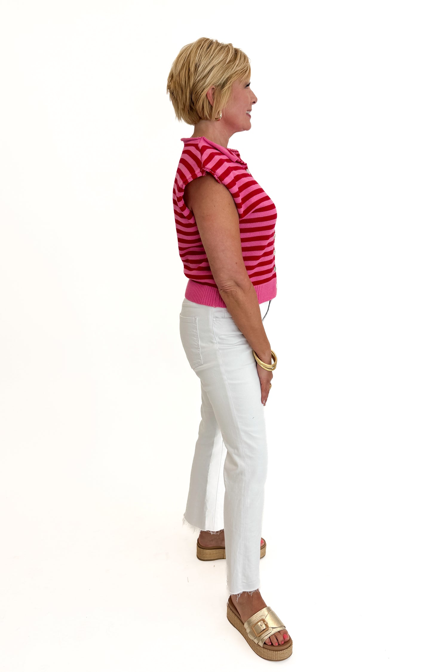 Side view of woman wearing Swagger Boutique’s Pink &amp; Red Striped Sleeveless Button Up Sweater by Entro with jeans against white studio background.