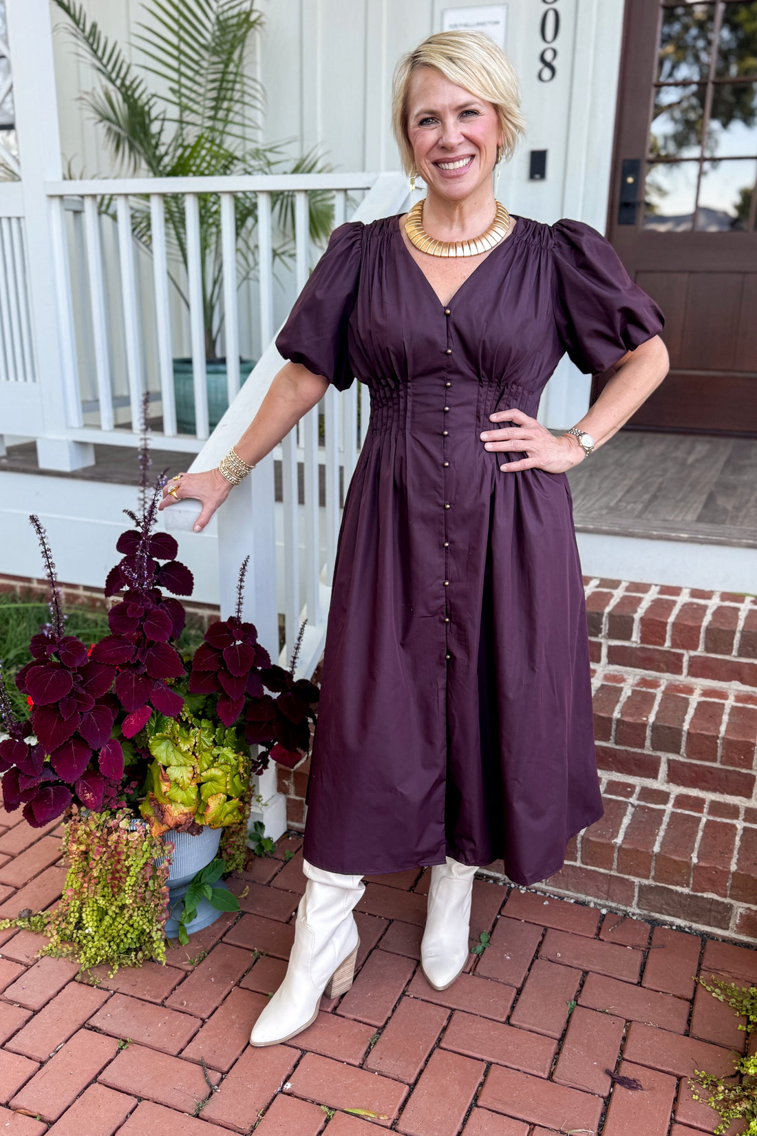 Front view of woman wearing Swagger Boutique’s Plum Pleated Waist Midi Dress by Entro with boots standing by a front porch outside.