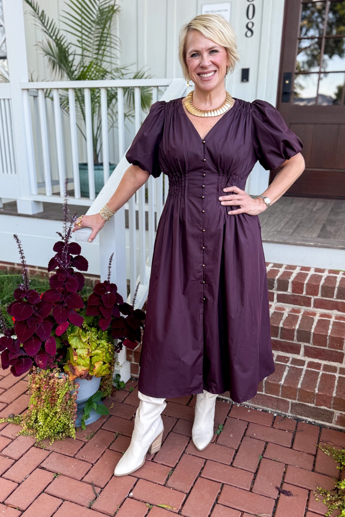 Front view of woman wearing Swagger Boutique’s Plum Pleated Waist Midi Dress by Entro with boots standing by a front porch outside.