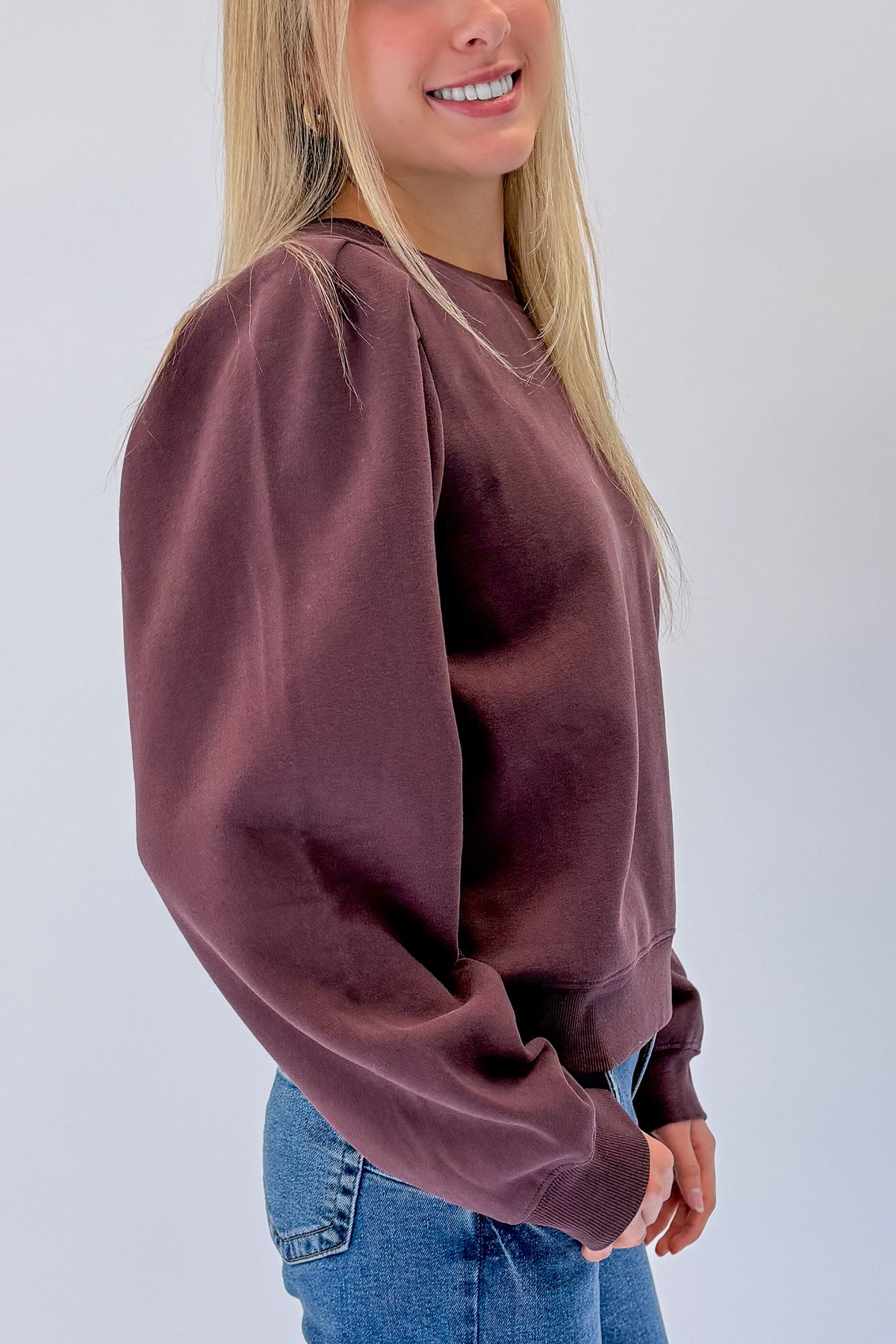 Side detail view of woman wearing Swagger Boutique’s Plum Puff Sleeve Sweatshirt with jeans against white studio background.