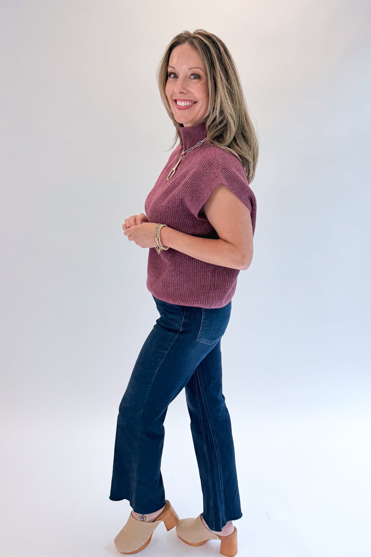 Side view of woman wearing Swagger Boutique’s Plum Sleeveless Mock Neck Sweater with jeans against white studio background.