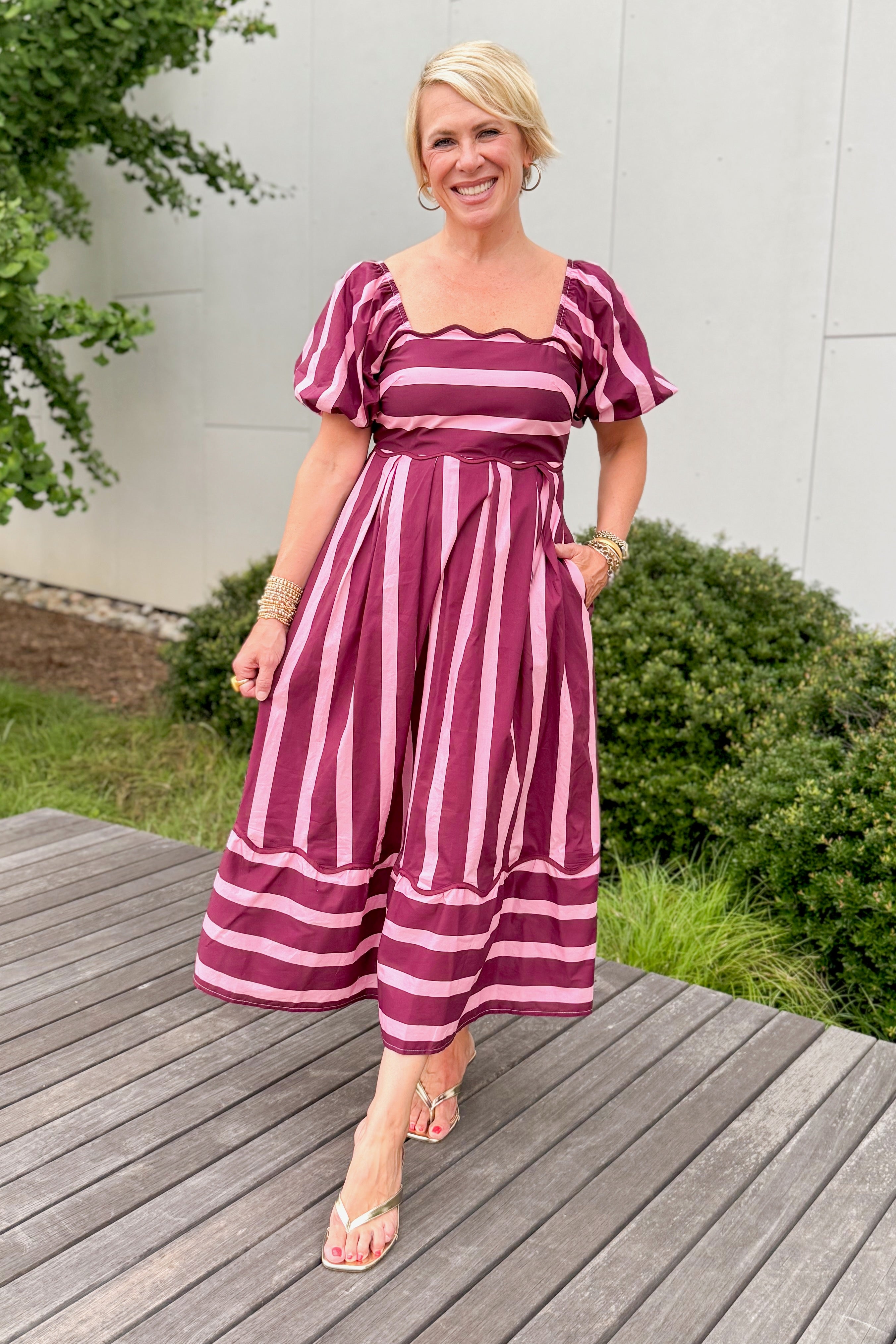 Front view of woman wearing Swagger Boutique’s Plum &amp; Pink Striped Midi Dress by Entro with sandals standing outside on a deck.