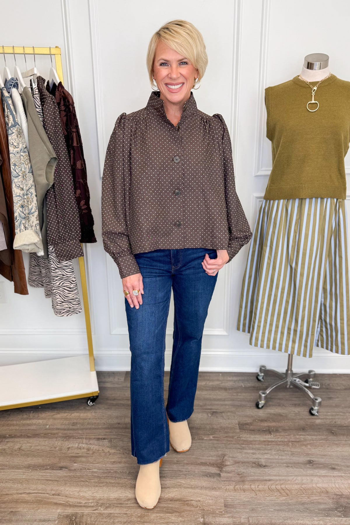 Front view of woman wearing Swagger Boutique’s Polka Dot Button Up Top with jeans with clothing hanging in the background.