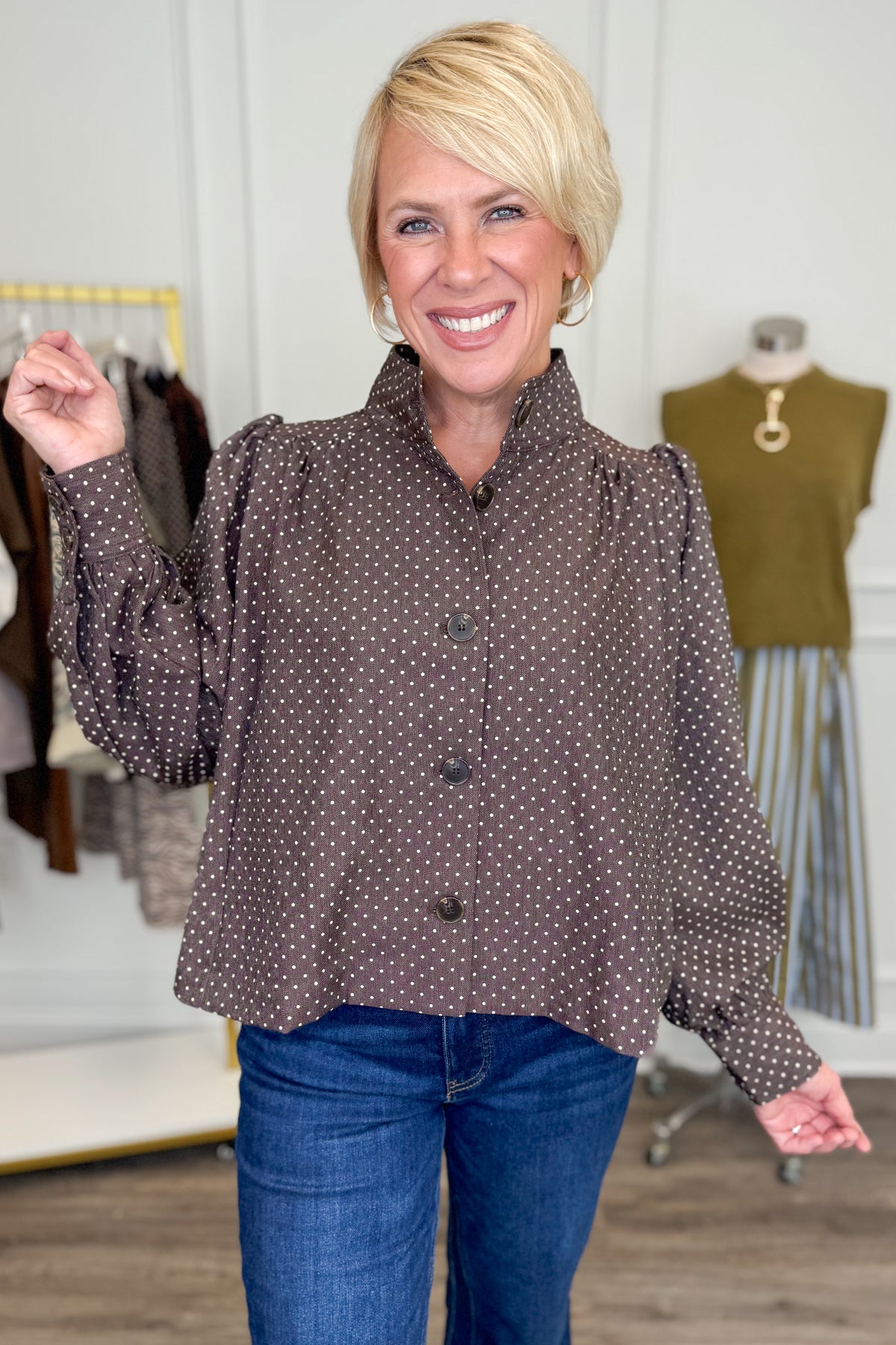 Front view of woman wearing Swagger Boutique’s Polka Dot Button Up Top with jeans with clothing hanging in the background.