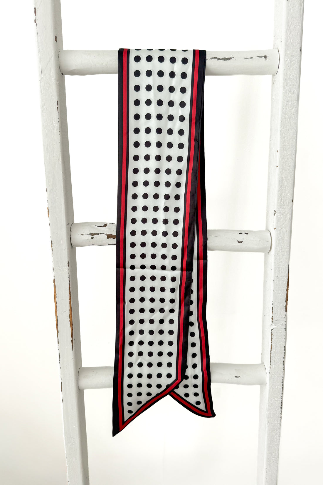 A long white scarf with black polka dots and red trim, draped over a white wooden ladder against a white background.