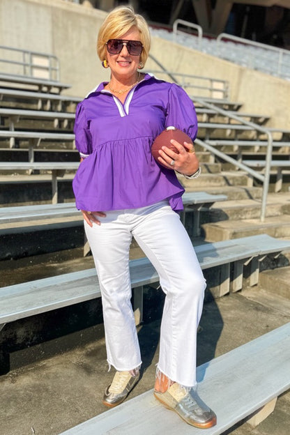 Front view of woman wearing Swagger Boutique’s Purple Collared Peplum Top with jeans outside at a stadium.